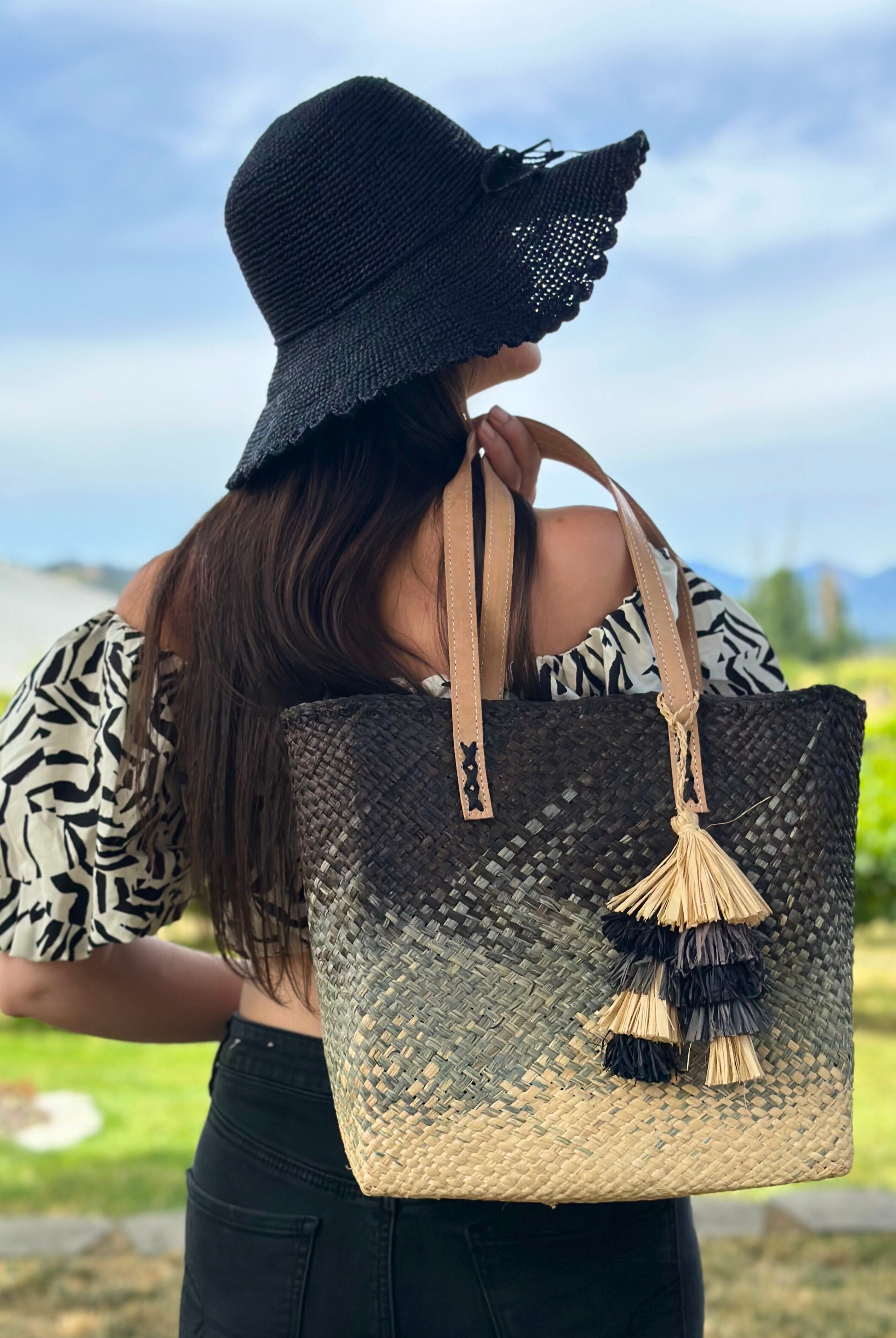 Model wearing Holden Ombre Black Straw Handbag Tassel Charm Embellishment handmade woven raffia dip dyed gradient of black, grey, and natural with matching multicolored, multi-tiered raffia tuft tassel with 4" Brim Genevieve Crochet Straw Sun Hats with Lace Weave Edge Black - Shebobo