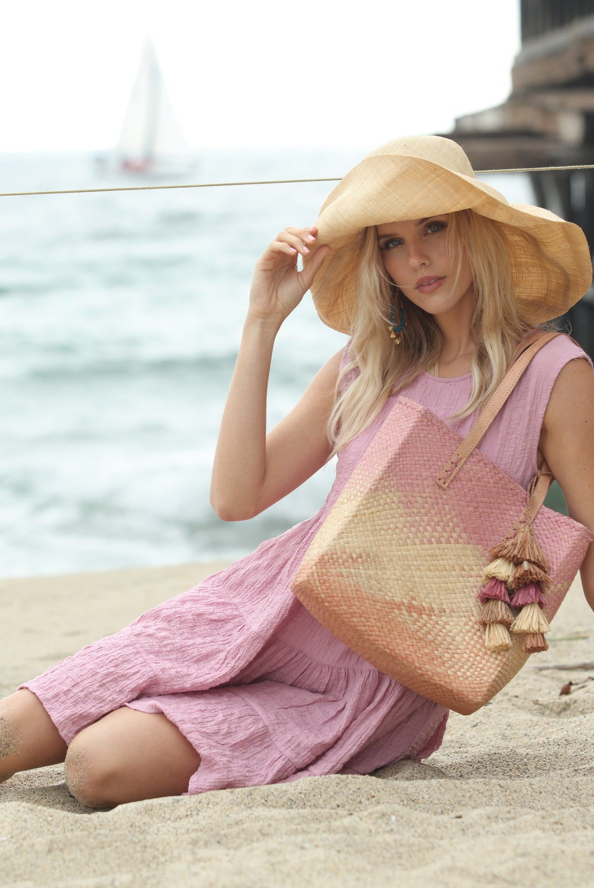 Model wearing Holden Ombre Wisteria Straw Handbag Tassel Charm Embellishment handmade woven raffia dip dyed gradient of wisteria lavender/lilac/light/purple, natural straw color, and blush orange/pink with matching multicolored, multi-tiered raffia tuft tassel - Shebobo (with 7" Wide Brim Natural Packable Straw Sun Hat)