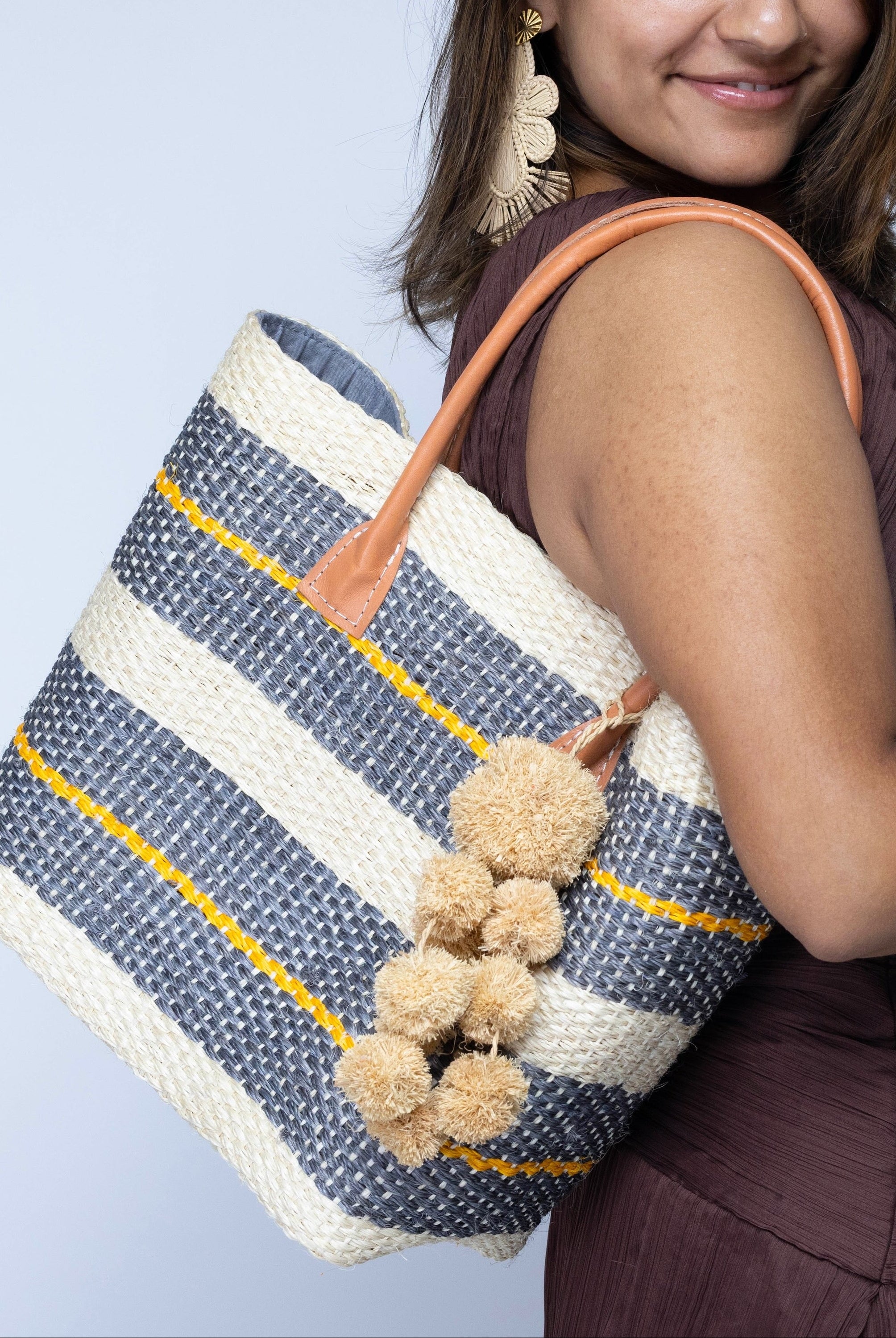 Model wearing Imperial Handbag natural sisal fiber in natural straw color, grey, and saffron yellow horizontal multi-width stripe pattern handmade woven basket with raffia waterfall pompom charm embellishment - Shebobo