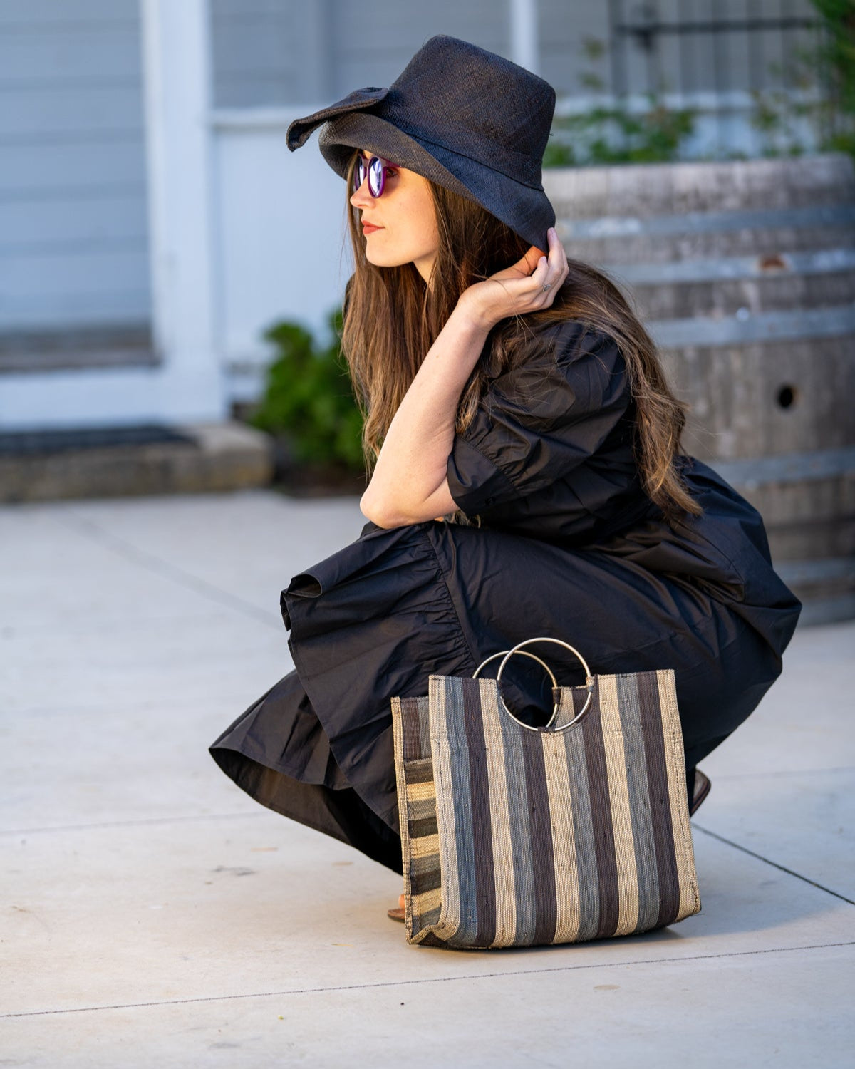 Model wearing Black Swirl JoJo Woven Straw Handbag with Metal Ring Handle handmade natural raffia palm fiber in a stripe pattern of natural straw color, grey, and black with matching woven thread texture and metallic circular metal ring handle - Shebobo (with 3" Brim Cara Bucket Straw Sun Hats with Big Bow)