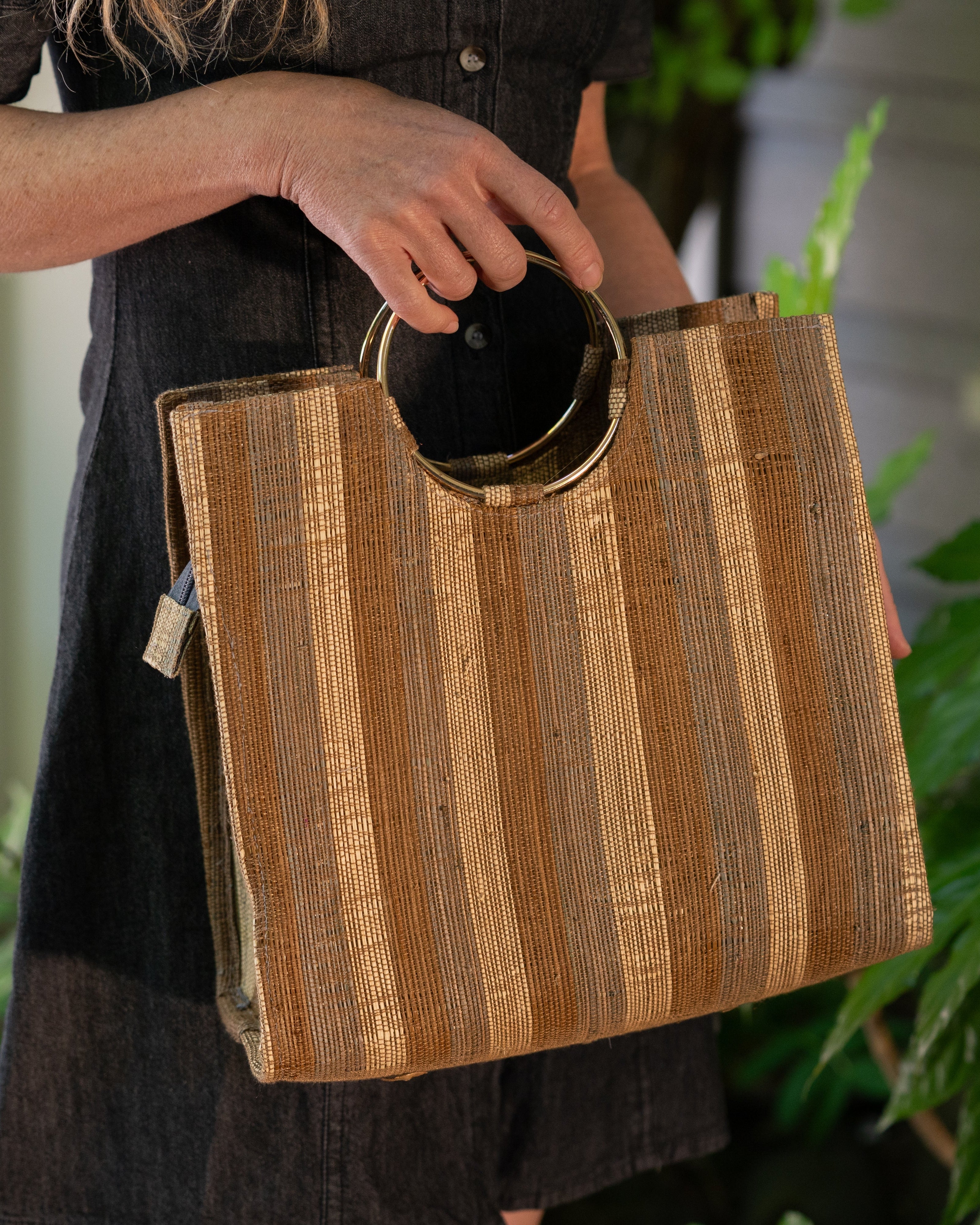 Model wearing Cinnamon Swirl JoJo Woven Straw Handbag with Metal Ring Handle handmade natural raffia palm fiber in a stripe pattern of natural straw color, grey, and Cinnamon tobacco/dark/brown with matching woven thread texture and metallic circular metal ring handle - Shebobo