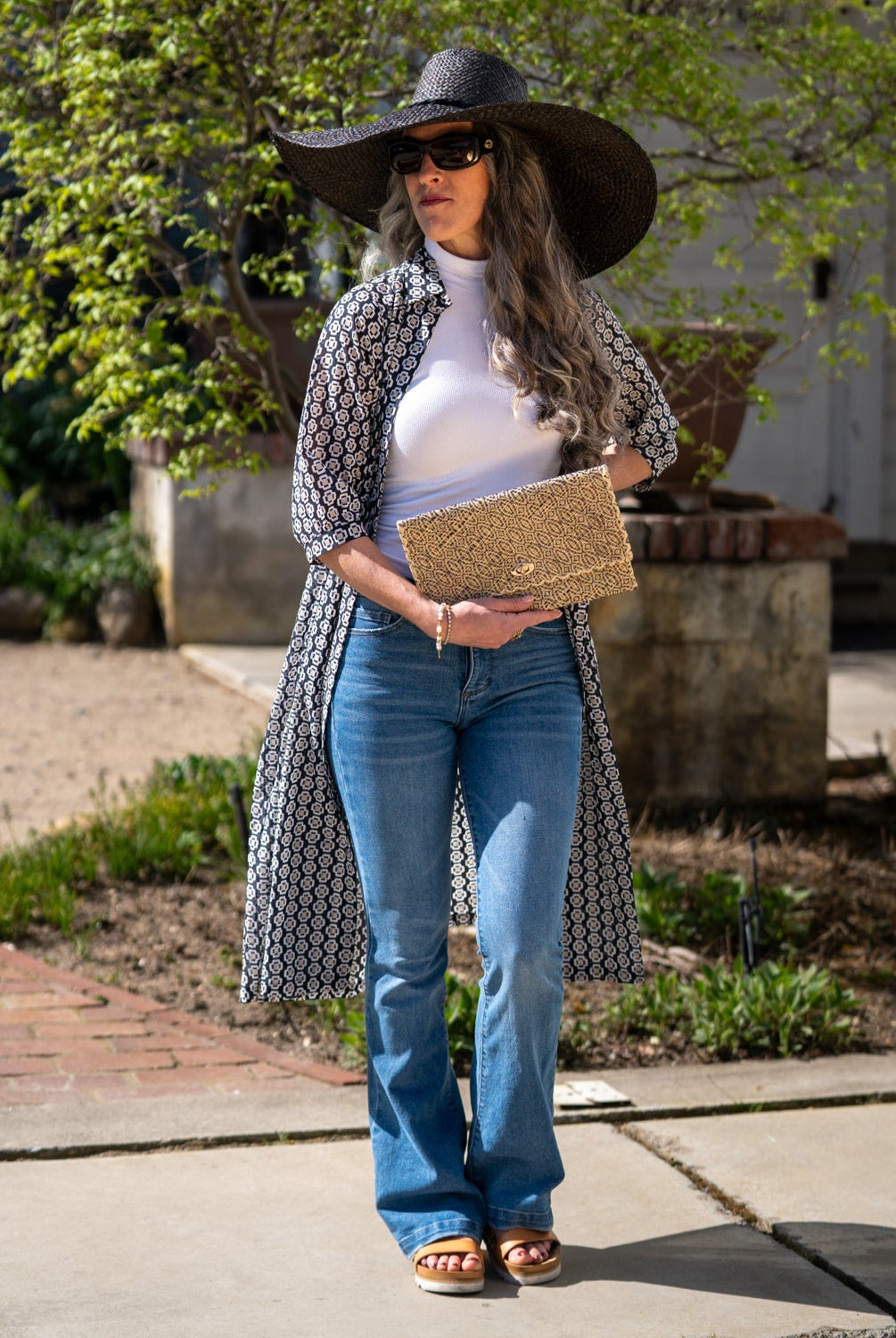 Model wearing Josephine Woven Straw Clutch Purse handmade natural raffia palm fiber in natural straw color woven with contrasting black thread stitching in a subtle diagonal stripe pattern consisting of bands of ovals and circles handbag with crosshatch stitch edging - Shebobo (with 7" Brim Natasha Woven XL Straw Sun Hats)