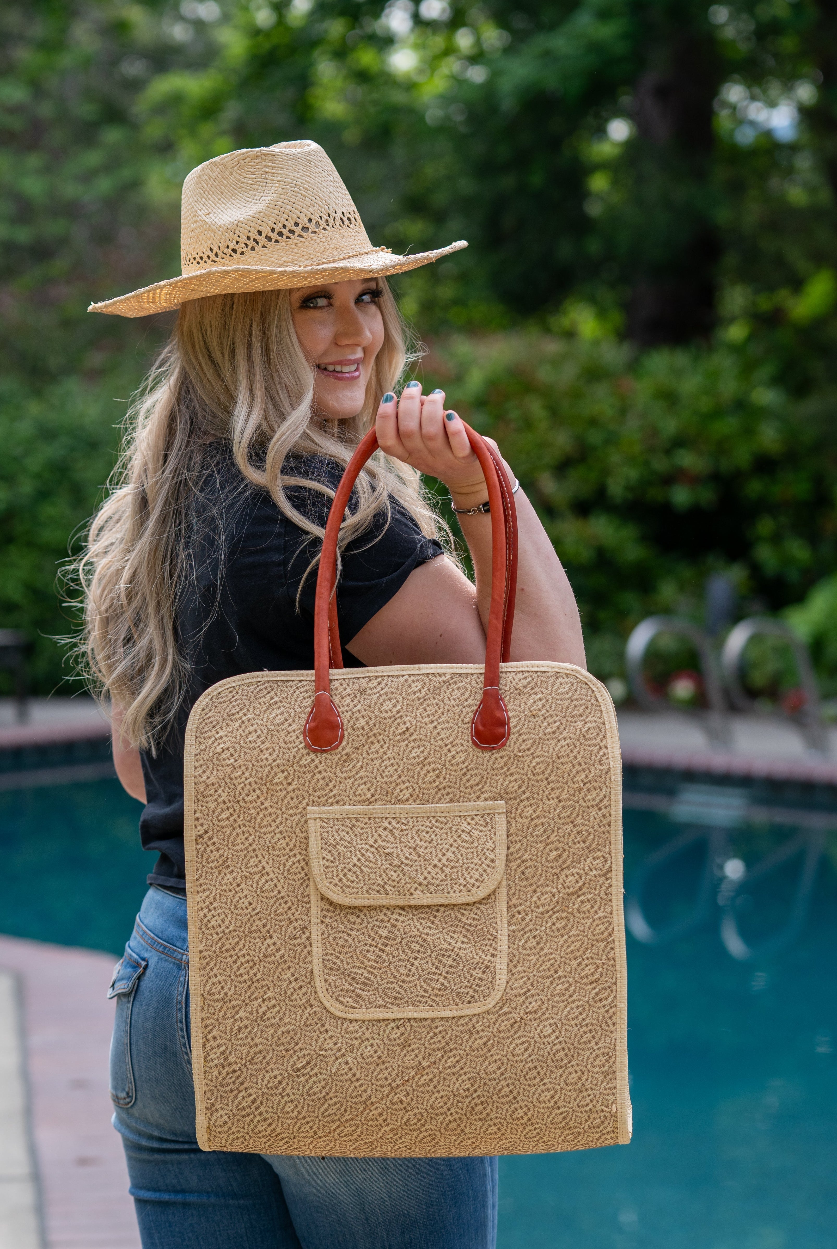 Model wearing Josephine 50's Inspired Woven Straw Handbag handmade natural raffia palm fiber in natural straw color woven with contrasting Cinnamon tobacco/dark/brown thread stitching in a subtle diagonal stripe pattern consisting of bands of ovals and circles with loomed raffia edging around the bag as well as the pocket and flap of the exterior pocket - Shebobo (with Macho Unisex Straw Cowboy Hats with Adjustable Wire Edge)
