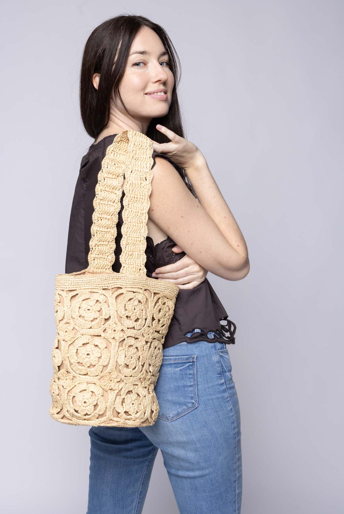 Model wearing Juliette Flower Crochet Raffia Straw Basket Bucket Handbag handmade natural raffia palm fiber in a solid hue of natural straw color woven into a geometric granny square floral cutout pattern with two size flowers around the circumference of the bag and a horizontal band of link texture around the top of the bag with scalloped edge woven single shoulder strap - Shebobo