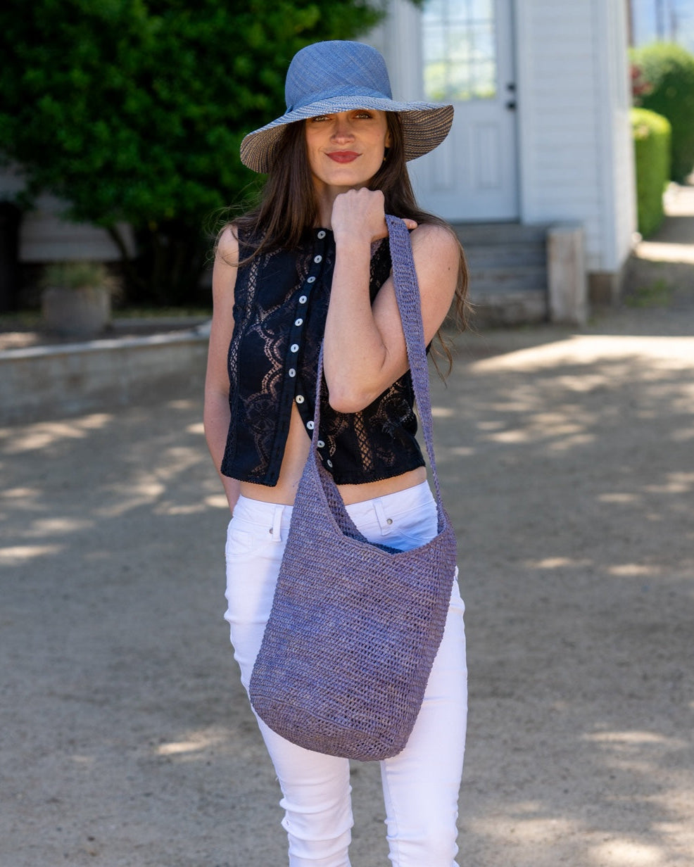 Model wearing Kiki Crochet Straw Crossbody Bag handmade natural raffia palm fiber in a crochet pattern of subtle linked bands in a solid hue of denim blue purse - Shebobo (with Marsea Marsea Two Tone Melange Straw Sun Hat)