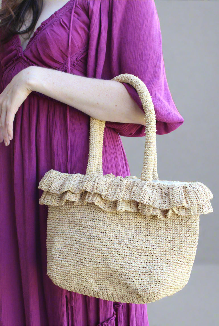 Model wearing Kiri Ruffle Crochet Straw Basket Handbag handmade crochet natural raffia palm fiber in a solid hue of natural straw color in subtle link texture with sweet double ruffle embellishment around the top of the bag and matching crochet handles beach bag shoulder bag - Shebobo