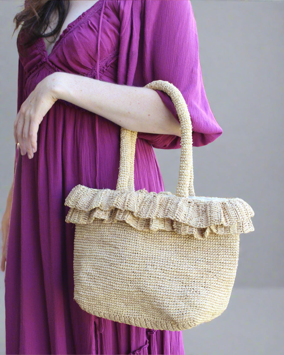 Model wearing Kiri Ruffle Crochet Straw Basket Handbag handmade crochet natural raffia palm fiber in a solid hue of natural straw color in subtle link texture with sweet double ruffle embellishment around the top of the bag and matching crochet handles beach bag shoulder bag - Shebobo