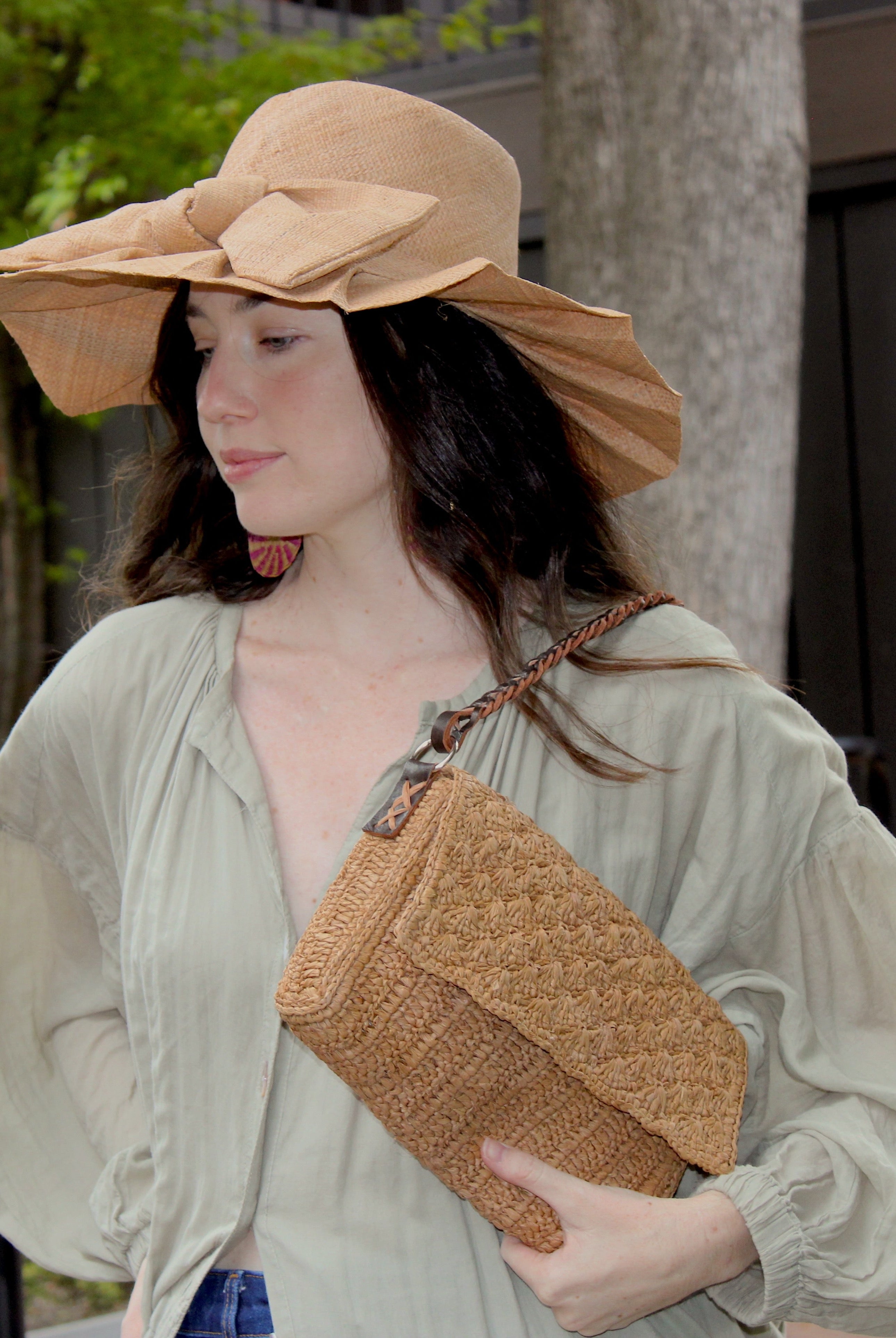 Model wearing Lilly 5" Brim Fan Straw Sun Hat with Big Bow Cappuccino handmade loomed natural raffia palm fiber in a solid hue of cappuccino tea/light/brown with pleated wide brim and oversized bow embellishment - Shebobo (with Anna Petal Pattern Crochet Straw Over Shoulder Envelope Clutch Purse)