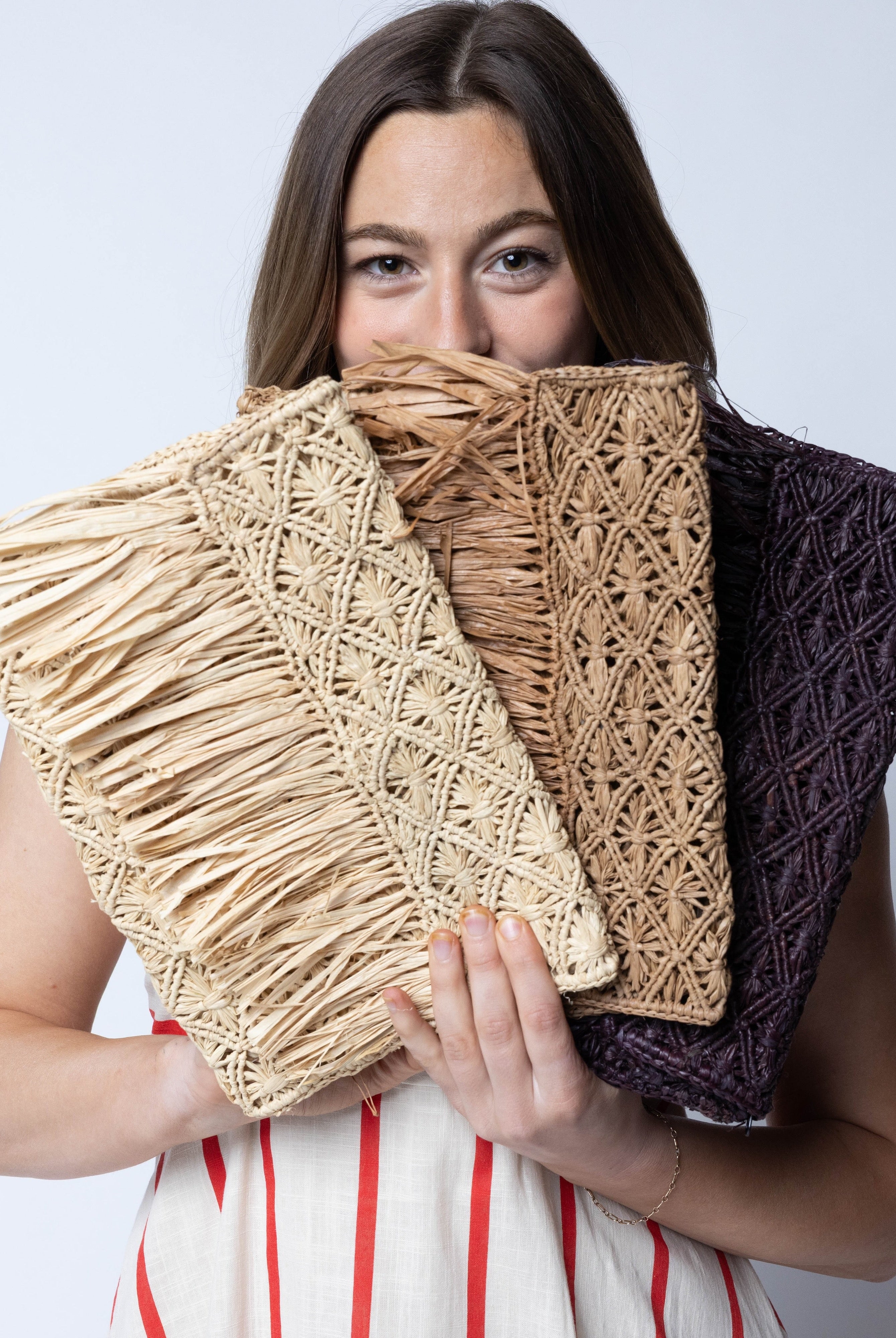Model wearing Multiple color options Luli Macrame Diamond and Crochet Straw Envelope Clutch Purse with Raw Fringe Trim Edge handmade macrame and crochet natural raffia palm fiber in a solid hue of Mocha, Natural, or Cappuccino in a diagonal geometric pattern of starburst or flower inside diamonds with hand brushed raw fringe trim edge along the top flap of the envelope design handbag - Shebobo