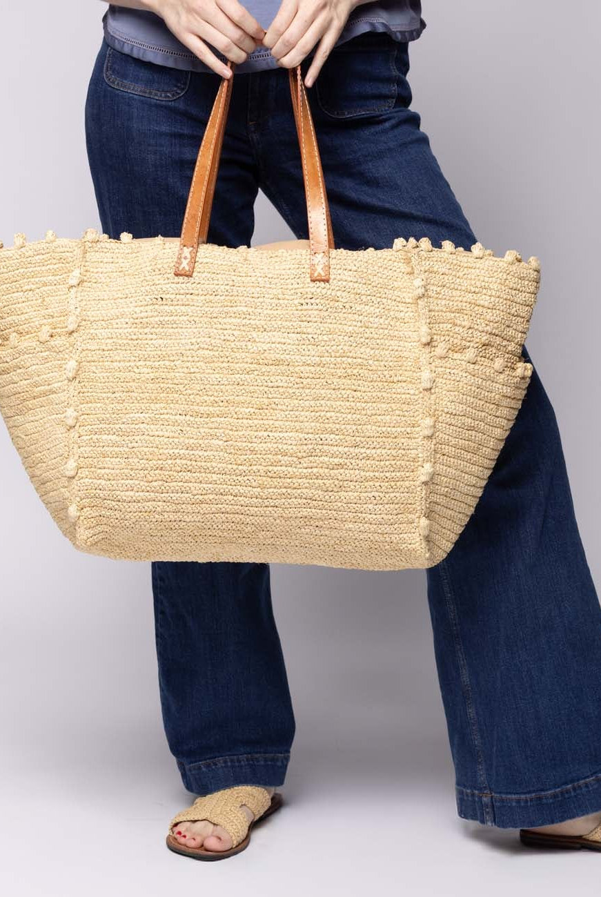 Model wearing Madeline Natural XL Crochet Raffia Straw Basket Shoulder Bag with Pockets handmade raffia palm fiber in a solid hue of natural straw color with crochet horizontal subtle band texture, round boucle nub edges, oversized external pockets, and leather handles - Shebobo