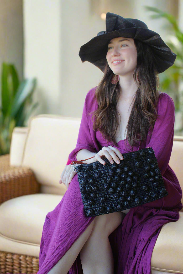Model wearing Marbella Crochet Straw Oversized Pouch Clutch Purse with Detachable Wristlet Strap handmade crochet natural raffia palm fiber in a solid hue of Black with subtle diamond boucle nub pattern protruding from horizontal bands of link texture small bag - Shebobo (Lilly 5" Brim Fan Pleated Straw Sun Hats with Big Bow)