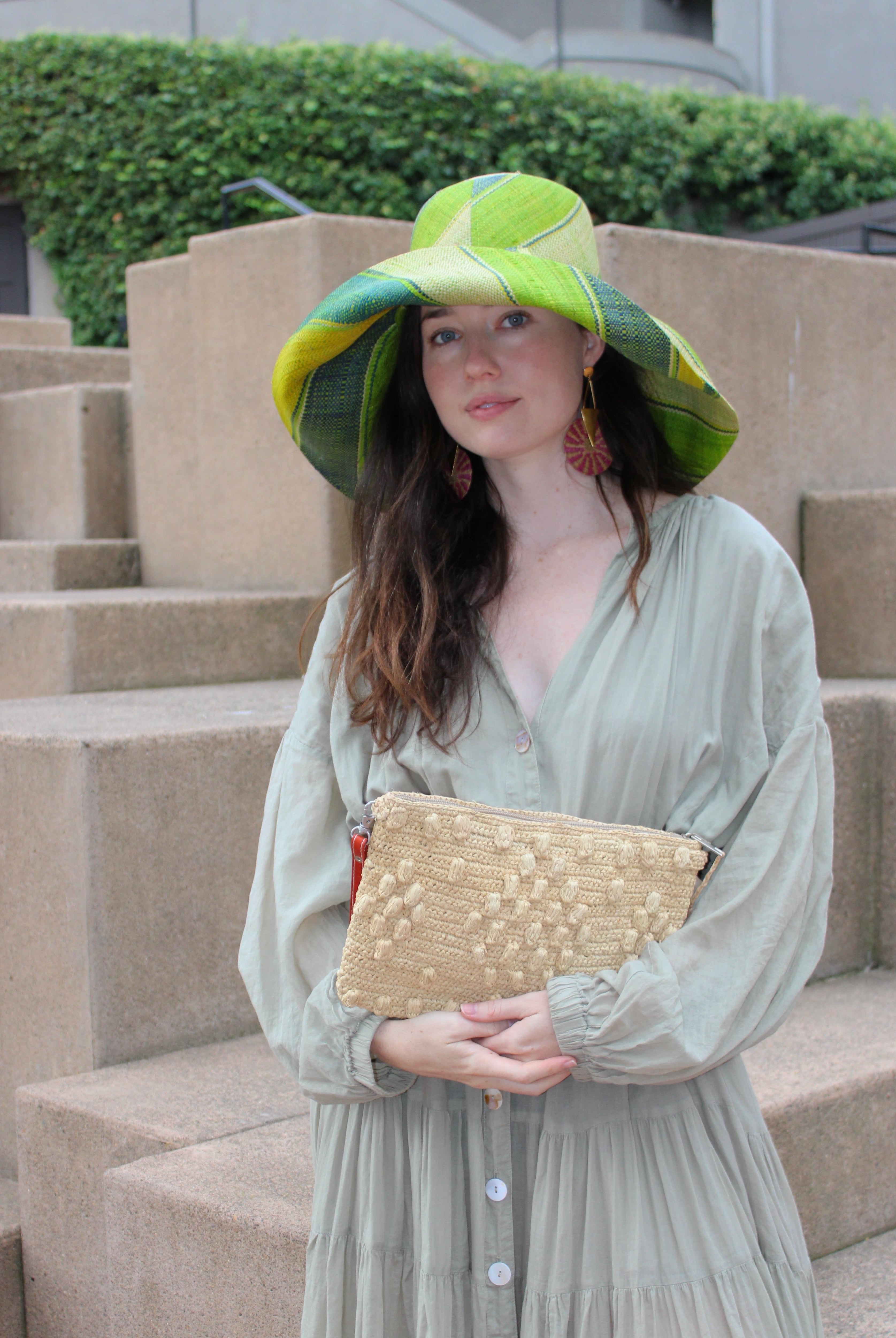 Model wearing Marbella Crochet Straw Oversized Pouch Clutch Purse with Detachable Wristlet Strap handmade crochet natural raffia palm fiber in a solid hue of Natural straw color with subtle diamond boucle nub pattern protruding from horizontal bands of link texture small bag - Shebobo (with Monica 5" & 7" Wide Brim Multicolor Stripe Pattern Packable Straw Sun Hats)