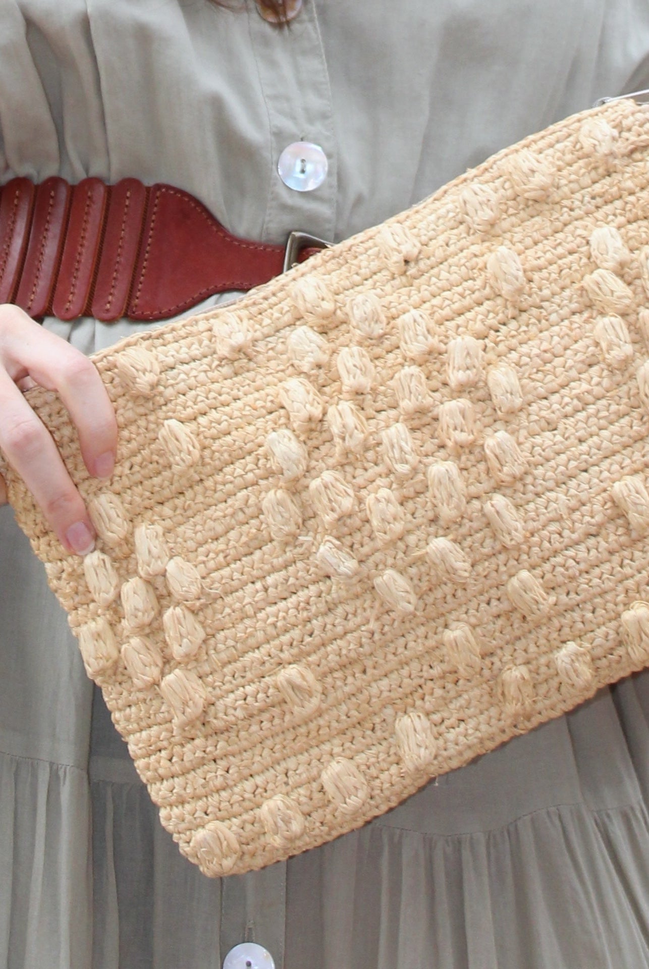 Model wearing Marbella Crochet Straw Oversized Pouch Clutch Purse with Detachable Wristlet Strap handmade crochet natural raffia palm fiber in a solid hue of Natural straw color with subtle diamond boucle nub pattern protruding from horizontal bands of link texture small bag - Shebobo