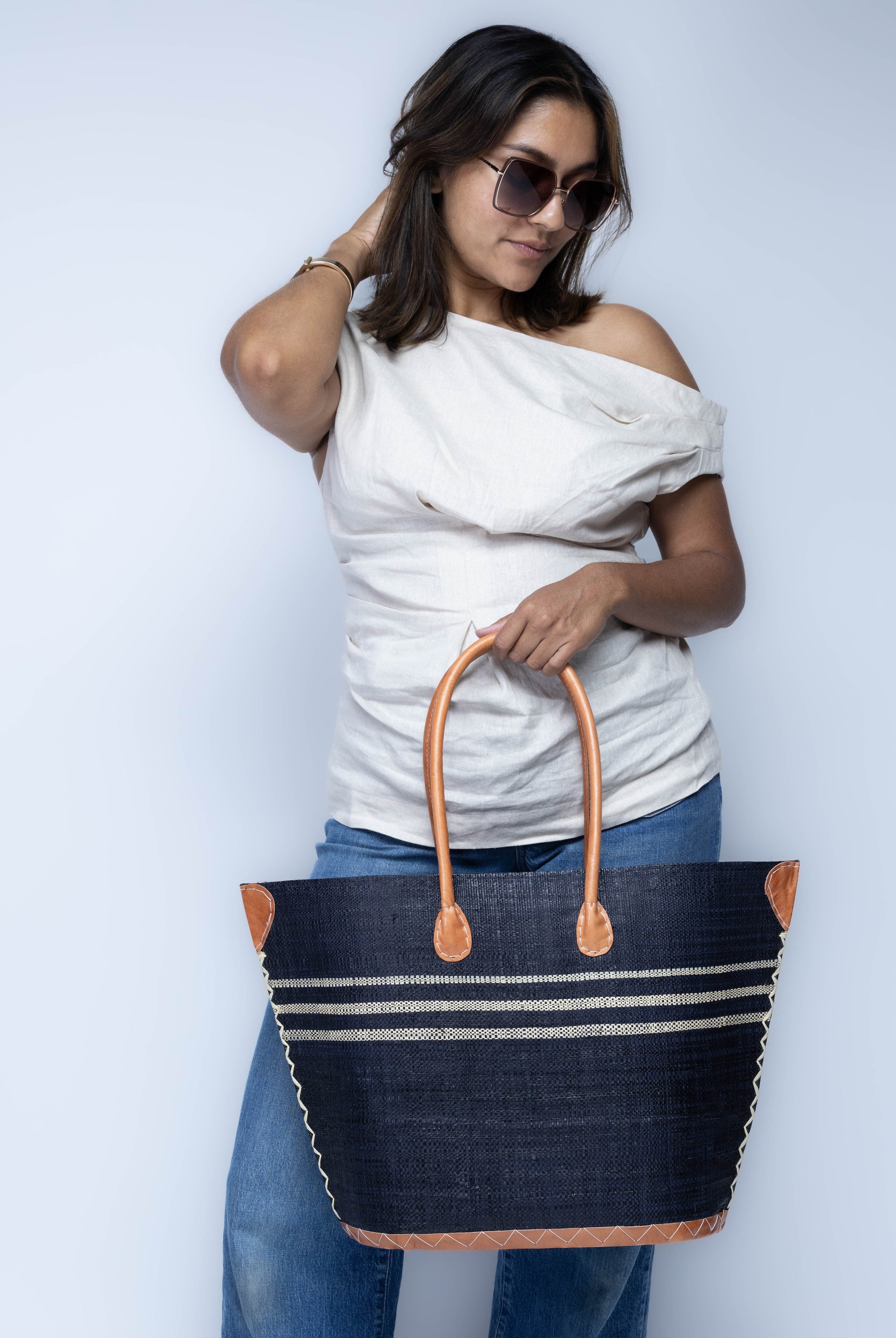 Model wearing Marina Racing Stripe Small Straw Tote Bag handmade loomed natural raffia palm fiber in a solid hue of black with three horizontal bands of natural straw color making a pinstripe pattern across the top third of the bag with matching cross-stitch edging, leather handles and accents beach bag handbag - Shebobo