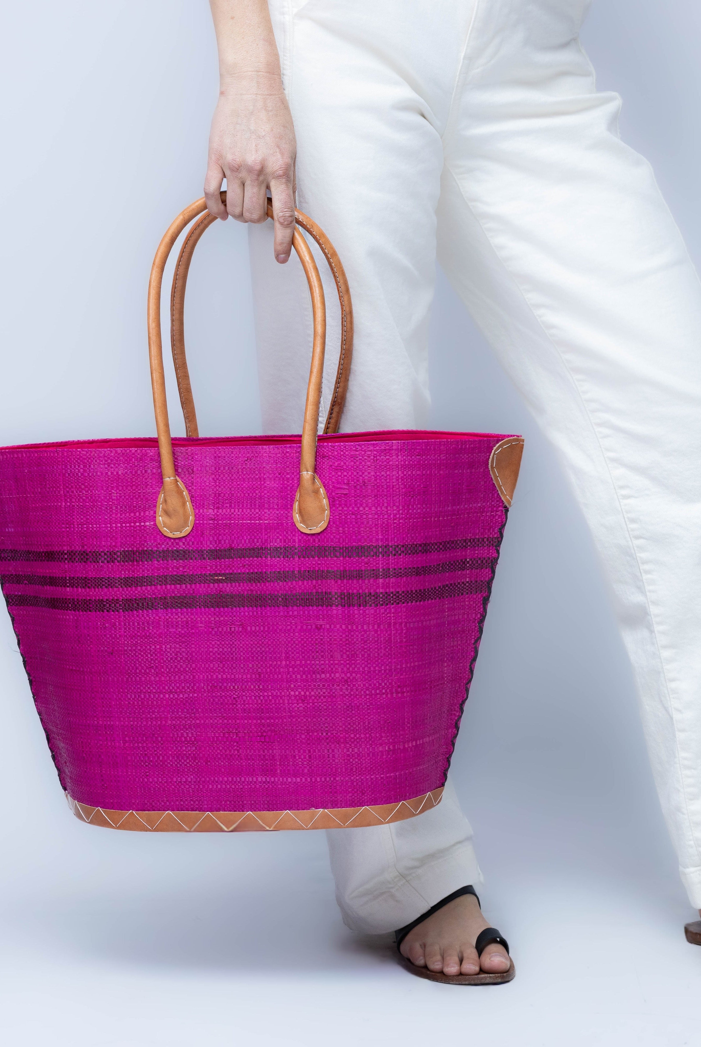 Model wearing Marina Racing Stripe Small Straw Tote Bag handmade loomed natural raffia palm fiber in a solid hue of Fuchsia hot/bright/barbie/pink with three horizontal bands of umberwine burgundy/dark/red/purple making a pinstripe pattern across the top third of the bag with matching cross-stitch edging, leather handles and accents beach bag handbag - Shebobo