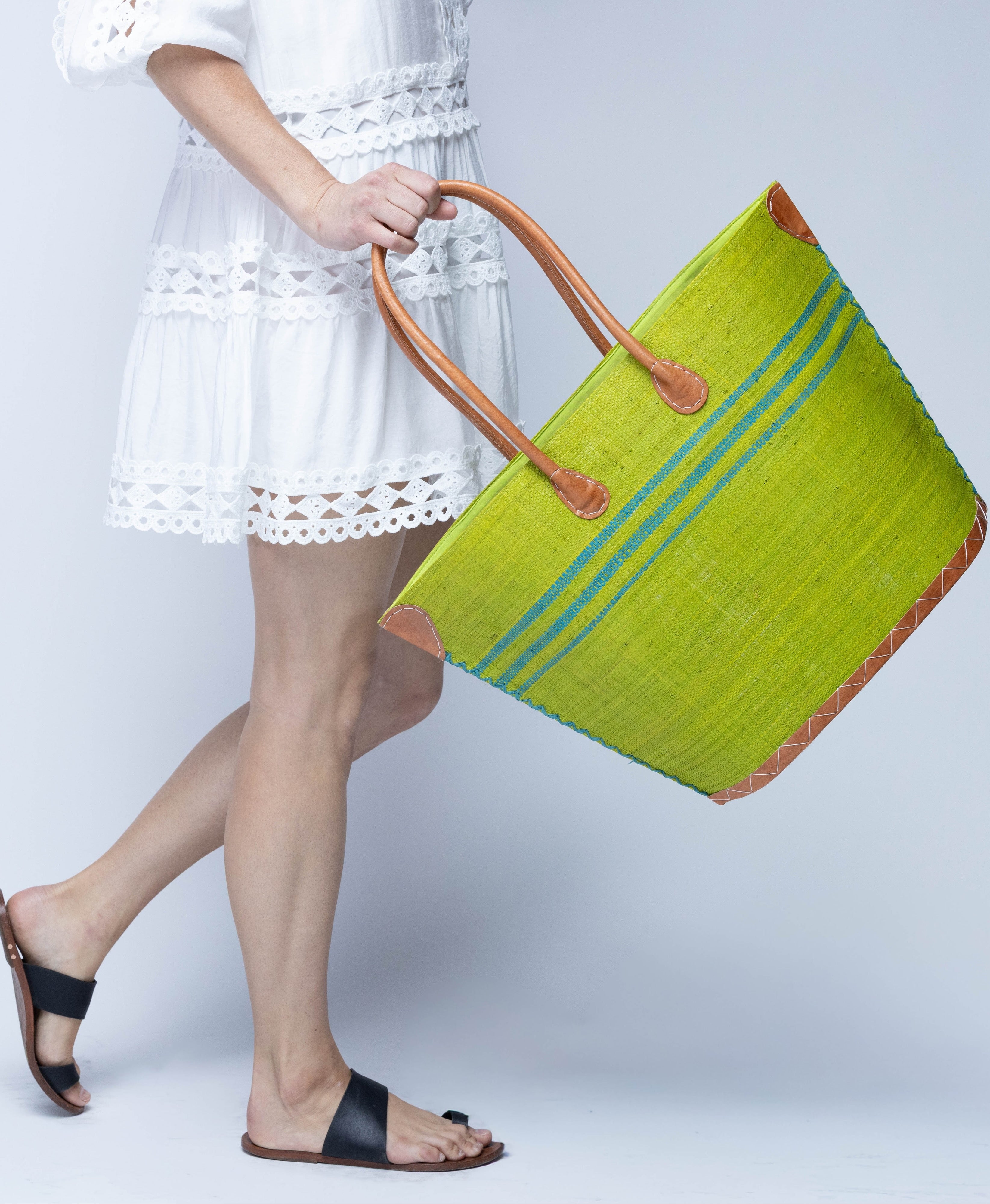 Model wearing Marina Racing Stripe Small Straw Tote Bag handmade loomed natural raffia palm fiber in a solid hue of midori bright/neon/lime/green with three horizontal bands of turquoise blue/green making a pinstripe pattern across the top third of the bag with matching cross-stitch edging, leather handles and accents beach bag handbag - Shebobo