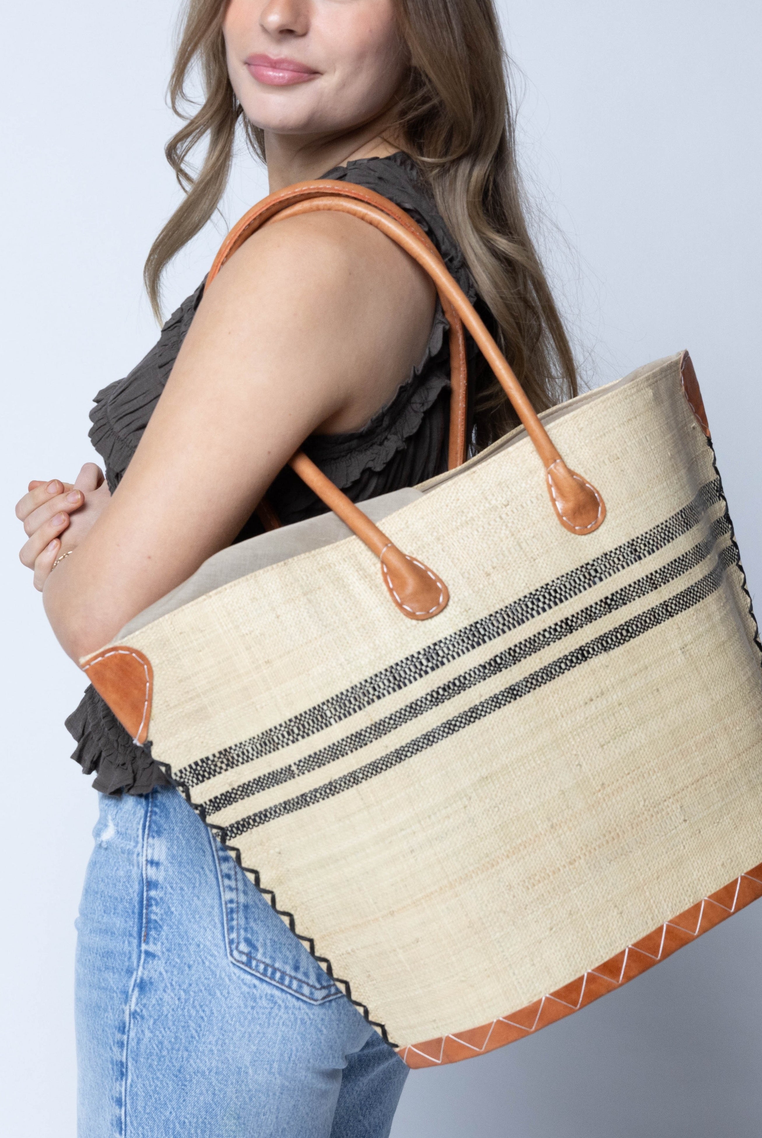 Model wearing Marina Racing Stripe Small Straw Tote Bag handmade loomed natural raffia palm fiber in a solid hue of natural straw color with three horizontal bands of black making a pinstripe pattern across the top third of the bag with matching cross-stitch edging, leather handles and accents beach bag handbag - Shebobo