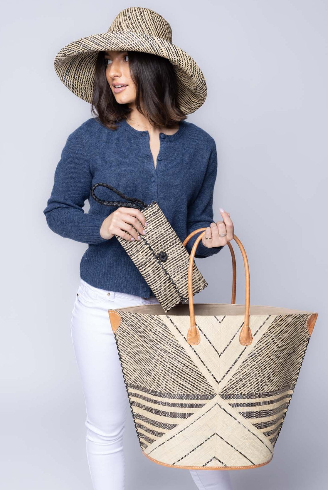 Model wearing Mariposa Large Straw Tote Bag handmade loomed natural raffia palm fiber in a multi paneled pattern design of pinstripe, melange, and wide stripe combinations geometrically radiating from the center point of the bag outwards in a two tone color combination of Natural straw color and black with a solid hue of color on the back of the bag plus leather handles and embellishments shoulder bag - Shebobo