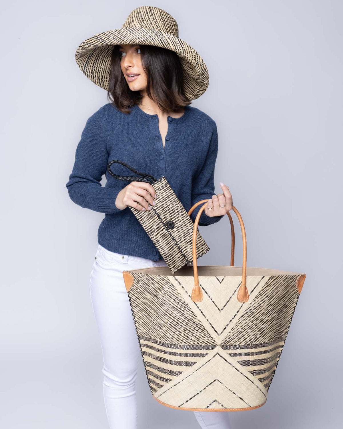 Model wearing Mariposa Large Straw Tote Bag handmade loomed natural raffia palm fiber in a multi paneled pattern design of pinstripe, melange, and wide stripe combinations geometrically radiating from the center point of the bag outwards in a two tone color combination of Natural straw color and black with a solid hue of color on the back of the bag plus leather handles and embellishments shoulder bag - Shebobo