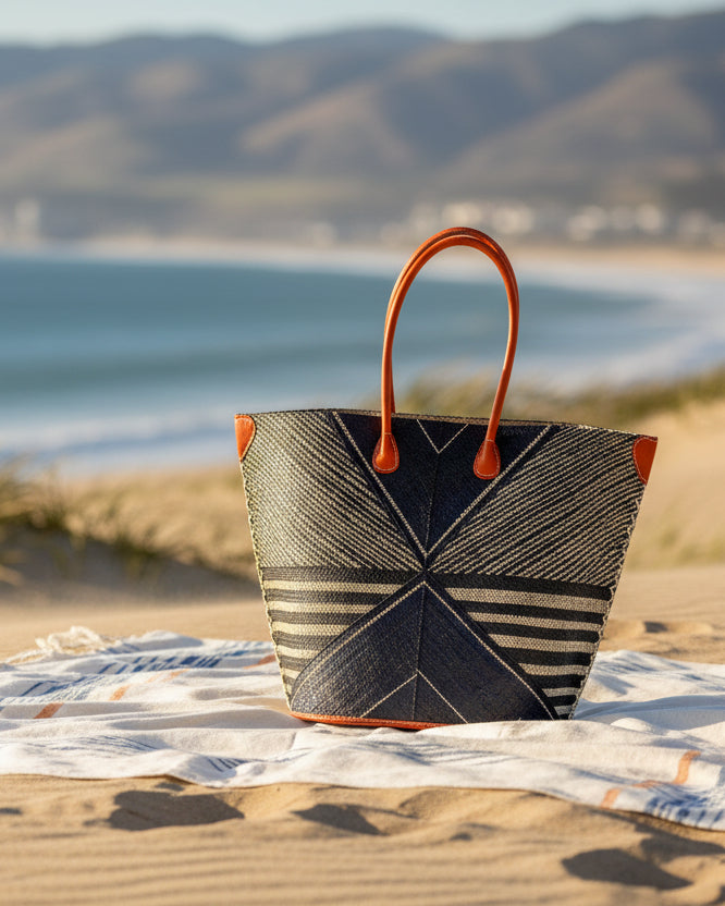 Lifestyle view Mariposa Large Straw Tote Bag handmade loomed natural raffia palm fiber in a multi paneled pattern design of pinstripe, melange, and wide stripe combinations geometrically radiating from the center point of the bag outwards in a two tone color combination of Black and natural straw color with a solid hue of color on the back of the bag plus leather handles and embellishments shoulder bag - Shebobo