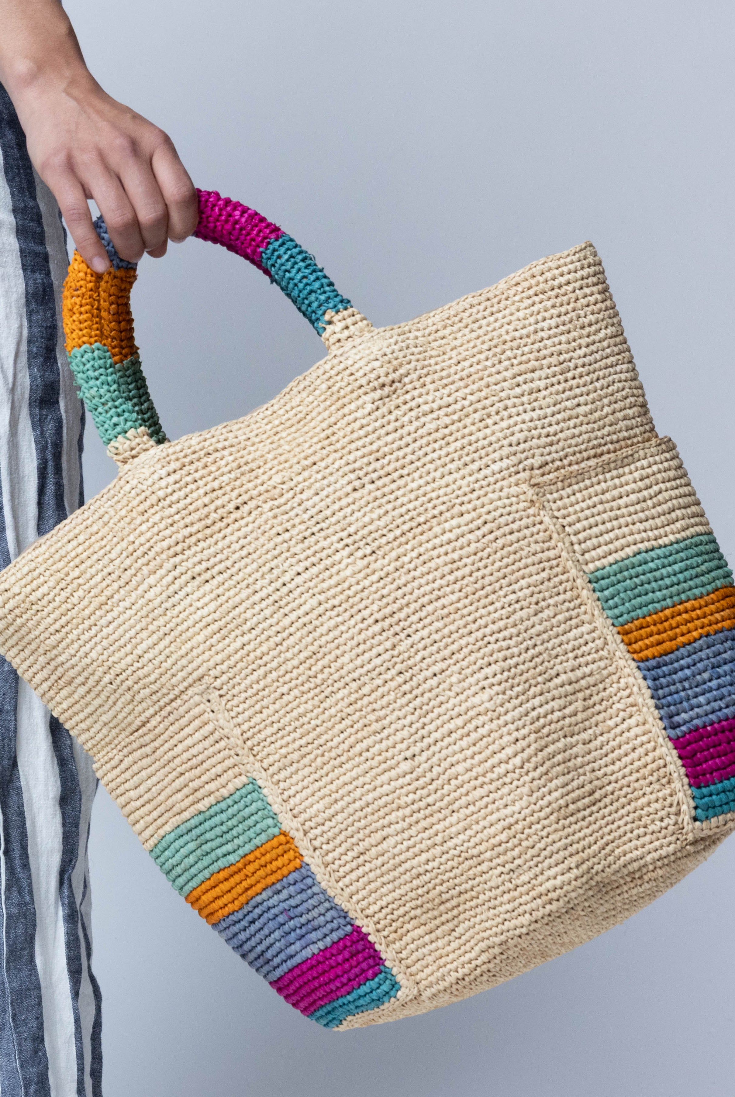 Model wearing Milos Caribbean Stripes Crochet Straw Basket Handbag with Pockets handmade natural raffia palm fiber in a solid hue of natural straw color on the body of the bag with multicolor wide stripe pattern of turquoise blue/green, fuchsia hot/bright/barbie/pink, denim retro/light/blue, saffron yellow/orange, and seafoam light/blue/green on both handles as well as two large external pockets tote bag - Shebobo