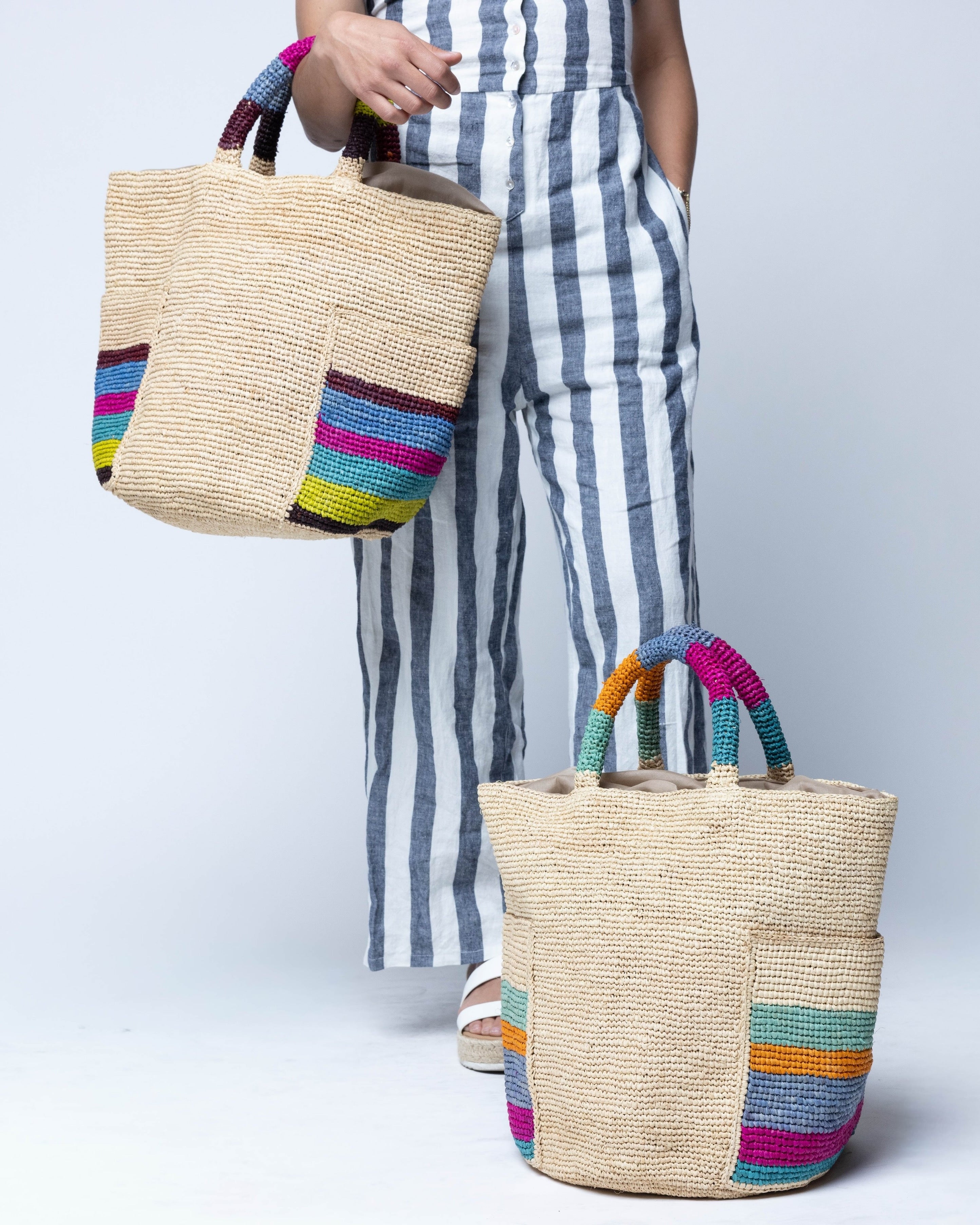 Model wearing Milos Caribbean and Riviera Stripes Crochet Straw Basket Handbag with Pockets handmade natural raffia palm fiber in a solid hue of natural straw color on the body of the bag with multicolor wide stripe pattern on both handles as well as two large external pockets tote bag - Shebobo