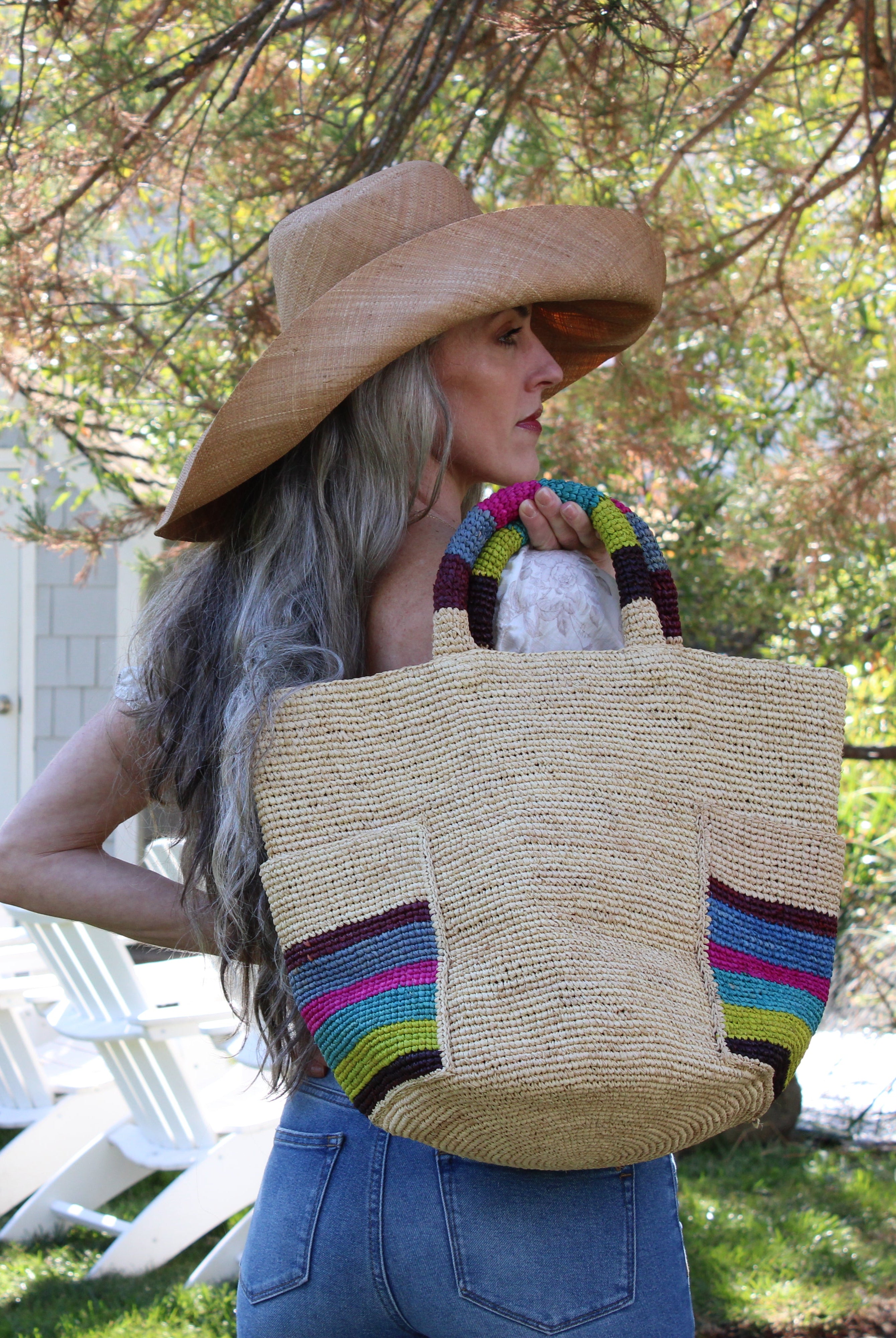 Model wearing Milos Riviera Stripes Crochet Straw Basket Handbag with Pockets handmade natural raffia palm fiber in a solid hue of natural straw color on the body of the bag with multicolor wide stripe pattern of umberwine bordeaux/claret/deep/dark/red/purple, denim retro/light/blue, fuchsia hot/bright/barbie/pink, teal dark/blue/green, midori lime/neon/green, and black on both handles as well as two large external pockets tote bag - Shebobo (with Audrey Wide Brim Big Bow Solid Color Packable Straw Sun Hat)