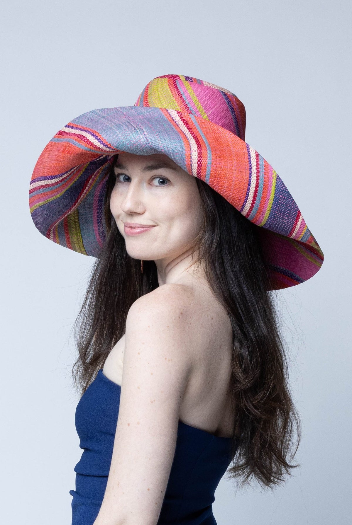 Model wearing Monica 7" Wide Brim Raspberry Swirl Multicolor Stripes Packable Straw Sun Hat Handmade loomed raffia in multi width bands of purple, light pink, coral orange/red, lime green, natural straw color, light blue, yellow, etc. create a swirl pattern - Shebobo