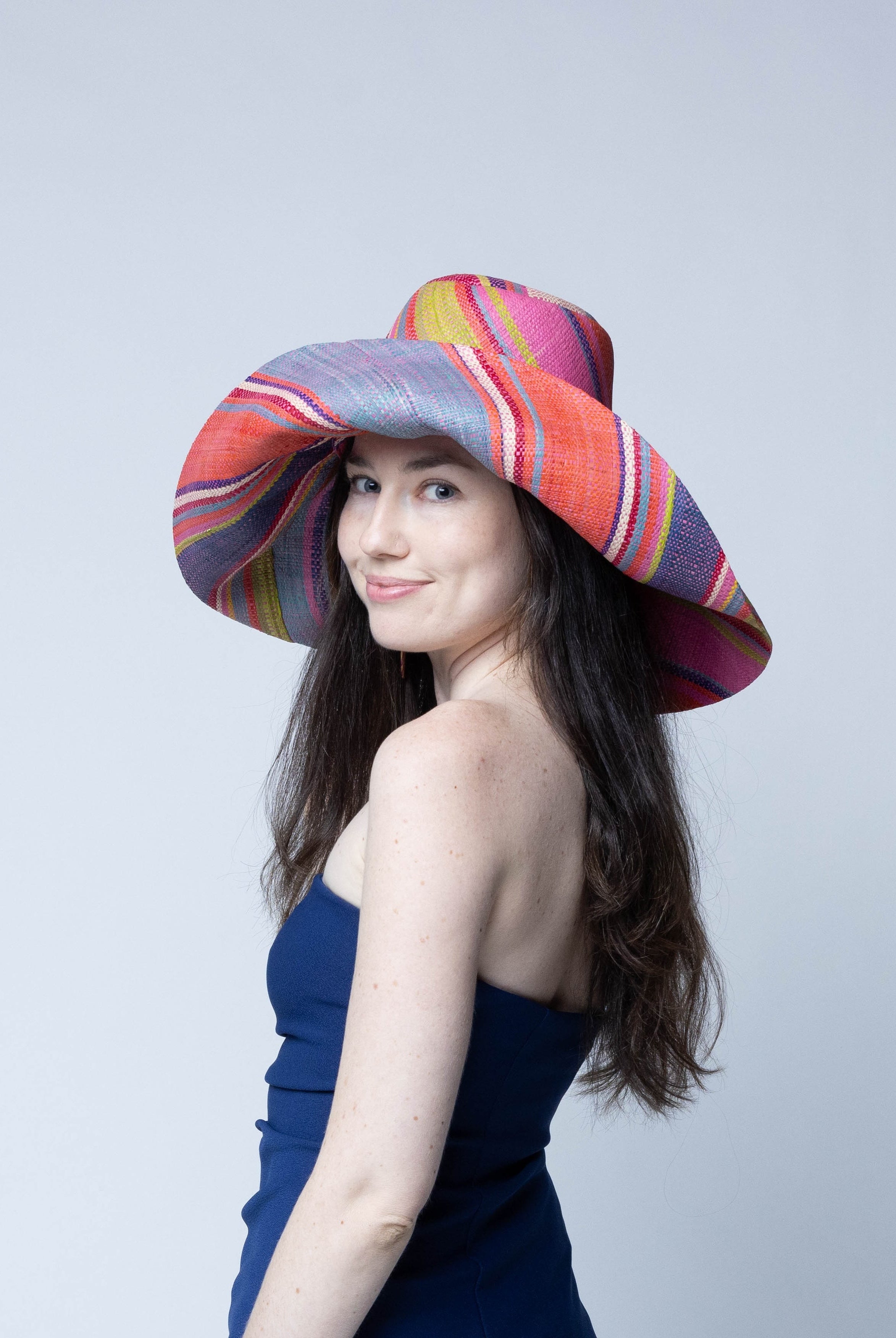 Model wearing Monica 7" Wide Brim Raspberry Swirl Multicolor Stripes Packable Straw Sun Hat Handmade loomed raffia in multi width bands of purple, light pink, coral orange/red, lime green, natural straw color, light blue, yellow, etc. create a swirl pattern - Shebobo