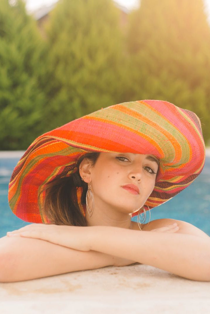 Model wearing Monica 7" Wide Brim Watermelon Swirl Multicolor Stripes Packable Straw Sun Hat Handmade loomed raffia in multi width bands of fuchsia pink, coral orange, red, yellow, black, lime green, etc. create a swirl pattern - Shebobo
