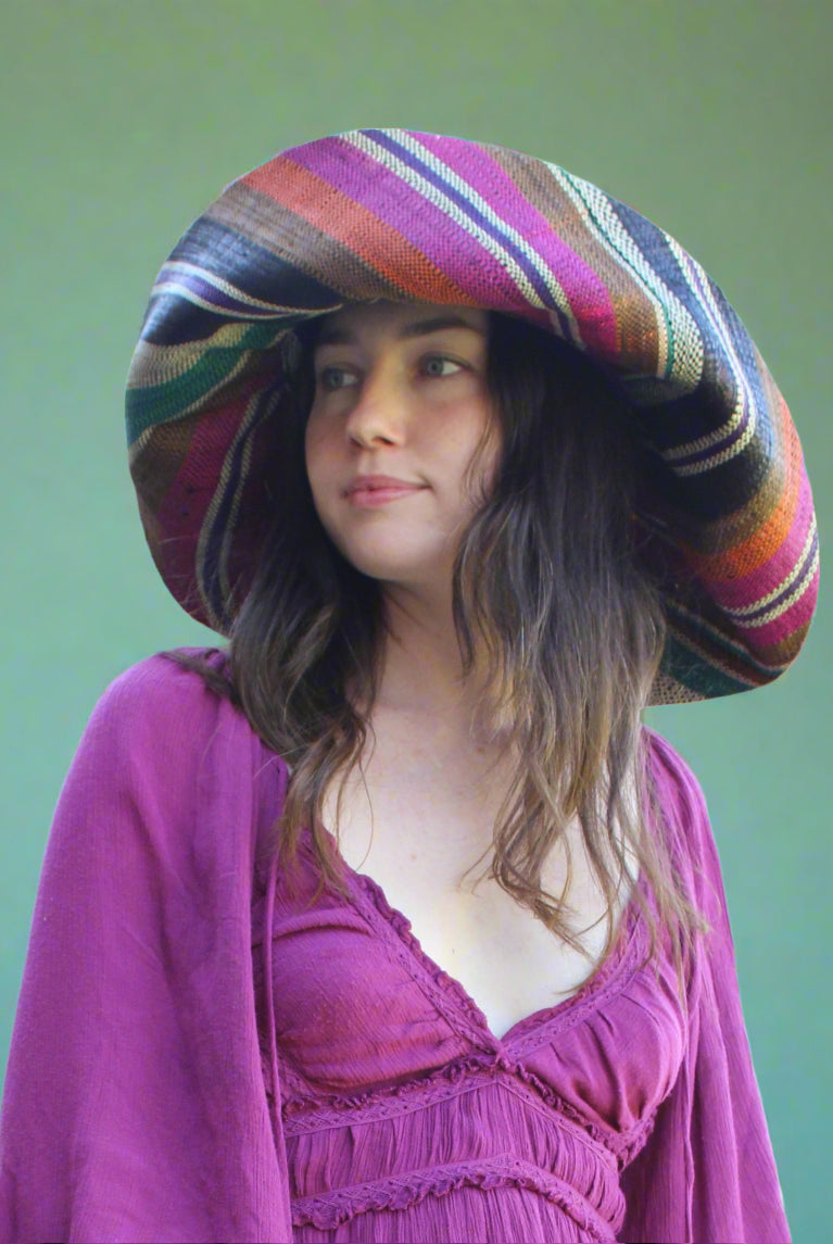 Model wearing Monica 7" Wide Brim Revel Swirl Multicolor Stripes Packable Straw Sun Hat Handmade loomed raffia in multi width bands of coral orange, fuchsia pink, teal blue/green, purple, cinnamon/tobacco/brown, black, natural straw color etc. create a swirl pattern - Shebobo