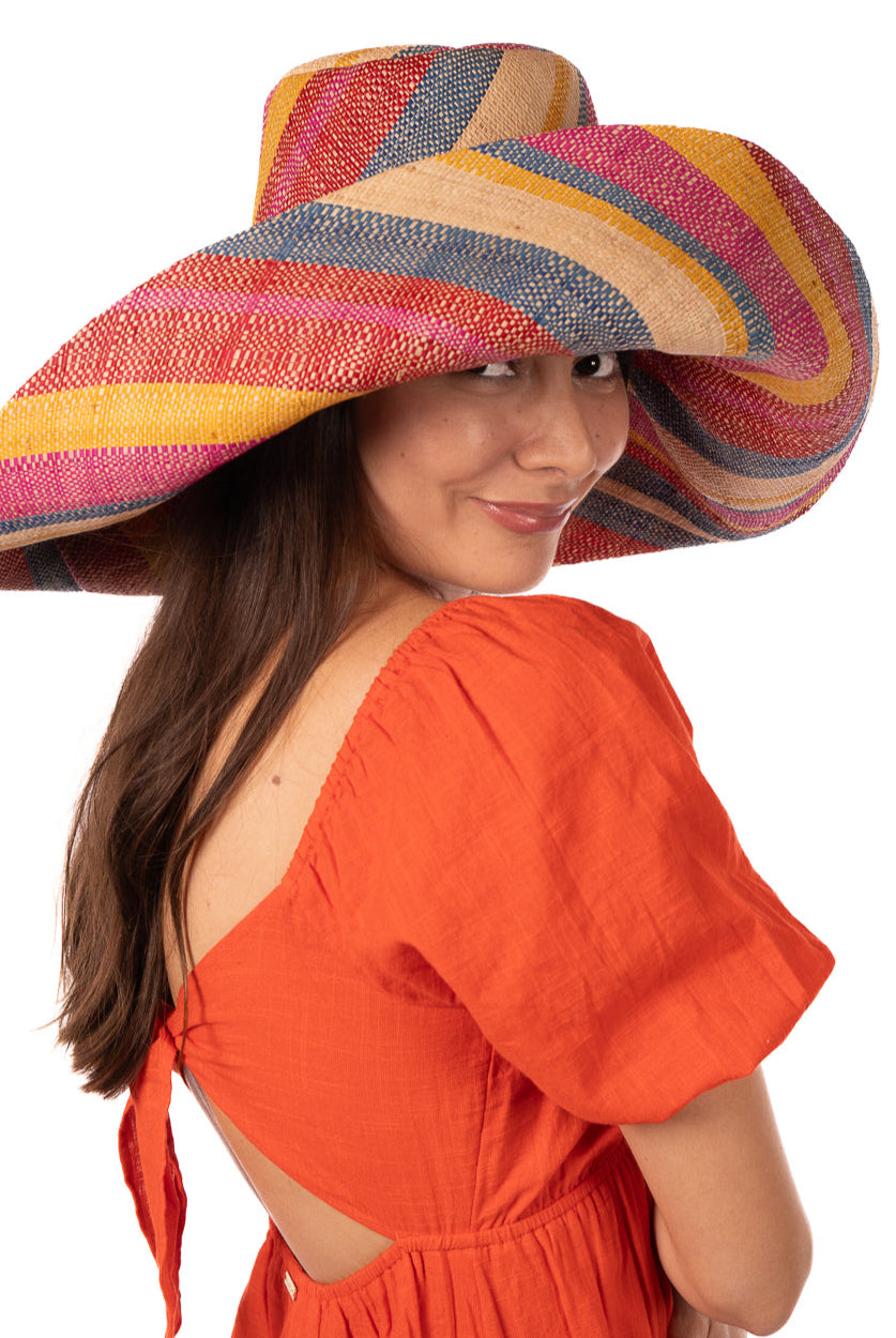 Model wearing Monica 7" Wide Brim Sassafras Swirl Multicolor Stripes Packable Straw Sun Hat Handmade loomed raffia in multi width bands of fuchsia pink, red, saffron yellow, navy blue, and natural straw color create a swirl pattern - Shebobo