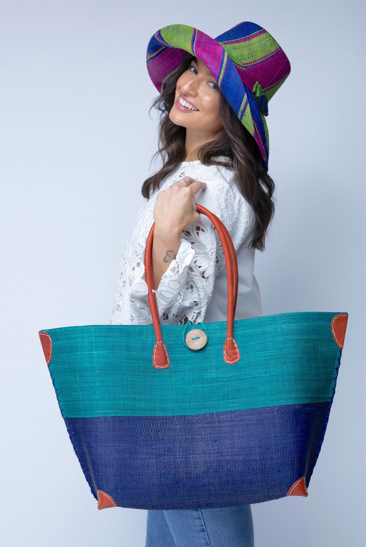Model wearing Monterey Two Tone Teal and Dusk Straw Tote Bag handmade loomed raffia fibers in a color block pattern of teal dark/blue/green colored upper half, and dusk dark/navy/blue lower half - Shebobo (with Monica 5" & 7" Wide Brim Multicolor Stripe Pattern Packable Straw Sun Hats)