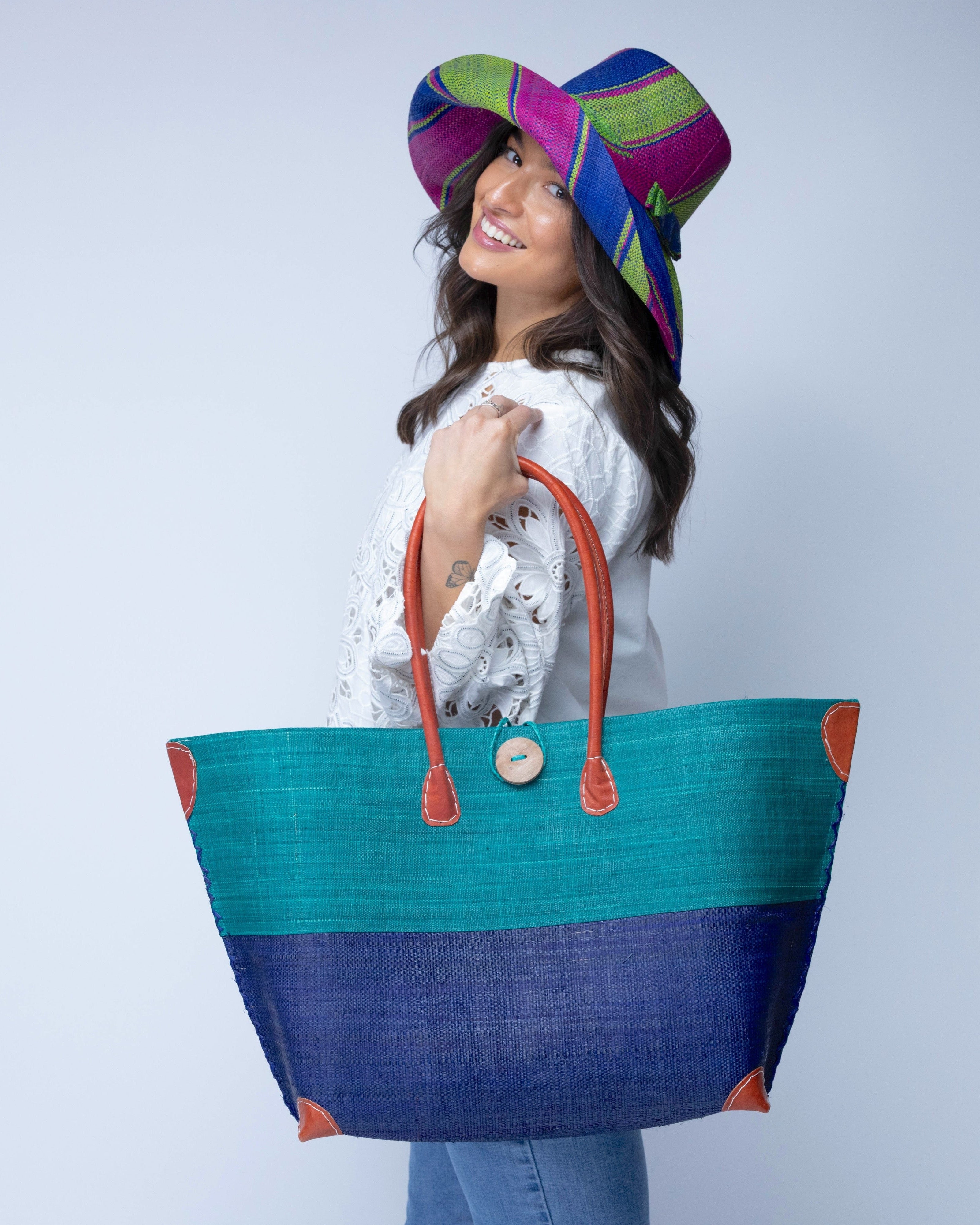 Model wearing Monterey Two Tone Teal and Dusk Straw Tote Bag handmade loomed raffia fibers in a color block pattern of teal dark/blue/green colored upper half, and dusk dark/navy/blue lower half - Shebobo (with Monica 5" & 7" Wide Brim Multicolor Stripe Pattern Packable Straw Sun Hats)
