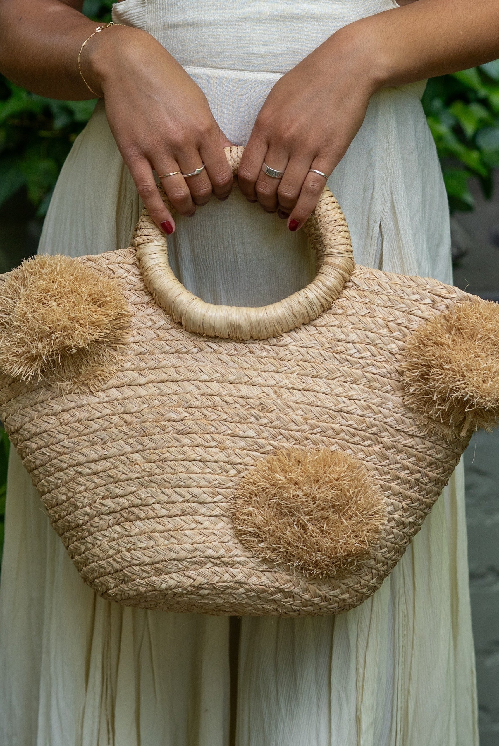 Model wearing Phi Phi Natural Braided Straw Basket with Oversized Raffia Poufs handmade raffia palm fiber braided into a solid hue of natural straw color with three randomly placed matching hand-brushed fringe poufs per side for embellishment with loop wrap top edging and crochet loop/braided handles bag handbag - Shebobo