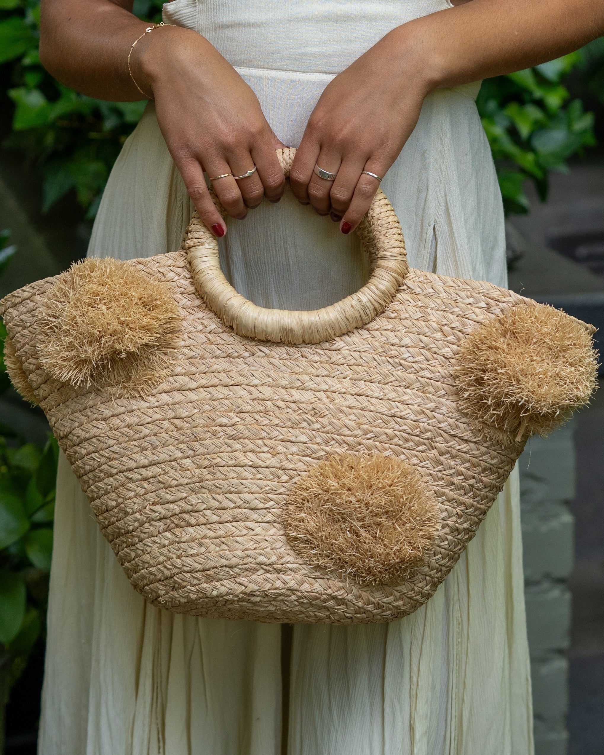Model wearing Phi Phi Natural Braided Straw Basket with Oversized Raffia Poufs handmade raffia palm fiber braided into a solid hue of natural straw color with three randomly placed matching hand-brushed fringe poufs per side for embellishment with loop wrap top edging and crochet loop/braided handles bag handbag - Shebobo