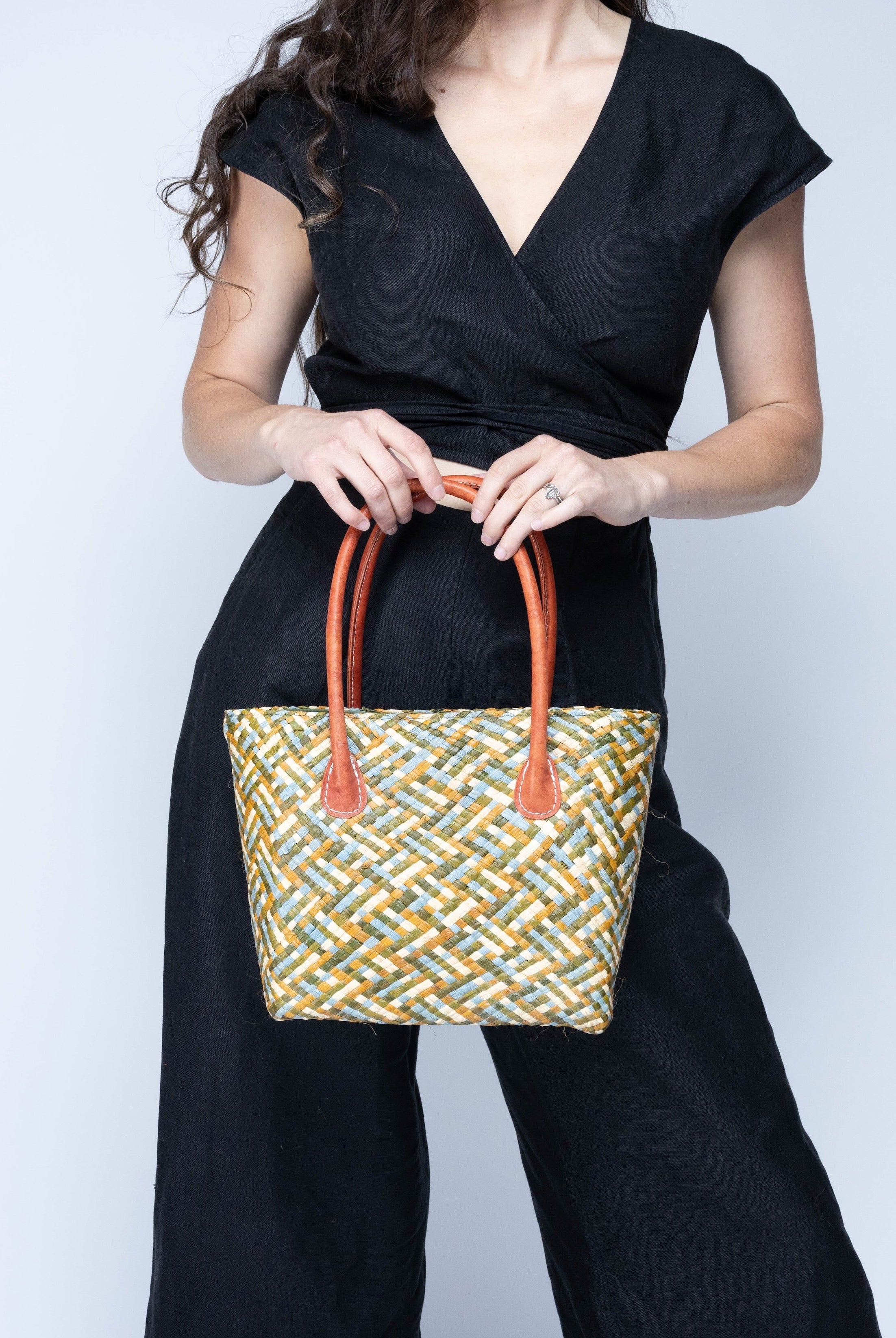 Model wearing Pianina Terra Multi Small Straw Basket Bag handmade from woven natural raffia palm fiber in a multicolor cross weave pattern of dark green, olive green, denim light/blue, and natural straw color handbag purse with leather handles - Shebobo