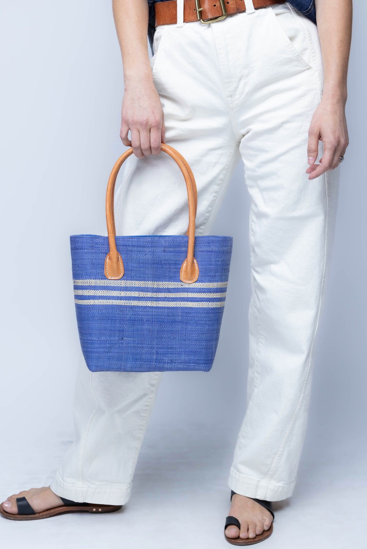 Model wearing Raja Racing Stripe Straw Basket Bag handmade loomed natural raffia palm fiber in a two tone multicolor design of Denim light/retro/soft/blue color with three horizontal bands of Natural straw color creating a racing stripe pattern placed under the base of the handles a third down the height of the bag - Shebobo