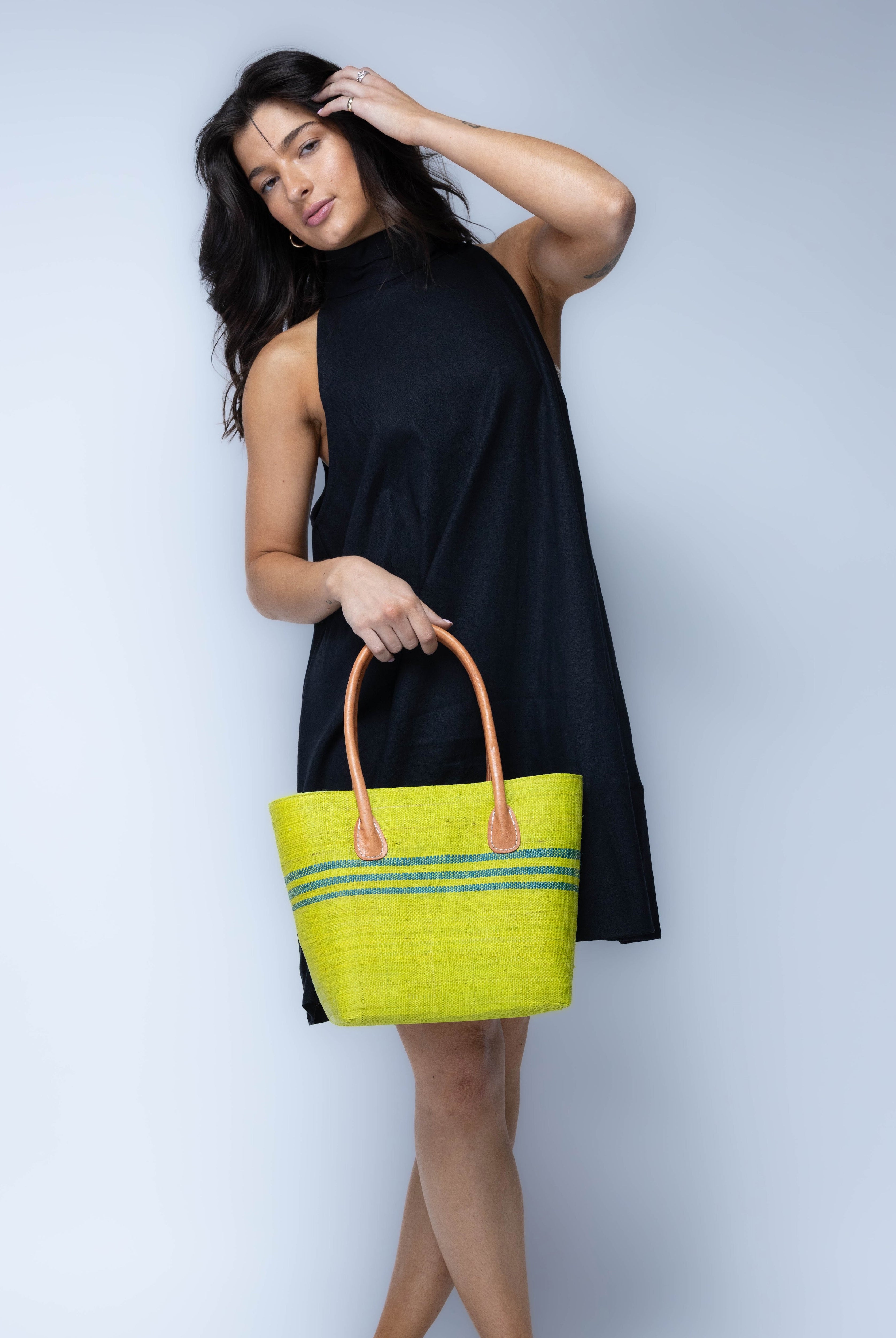Model wearing Raja Racing Stripe Straw Basket Bag handmade loomed natural raffia palm fiber in a two tone multicolor design of Midori lime/bright/neon/green color with three horizontal bands of Turquoise blue/green creating a racing stripe pattern placed under the base of the handles a third down the height of the bag - Shebobo