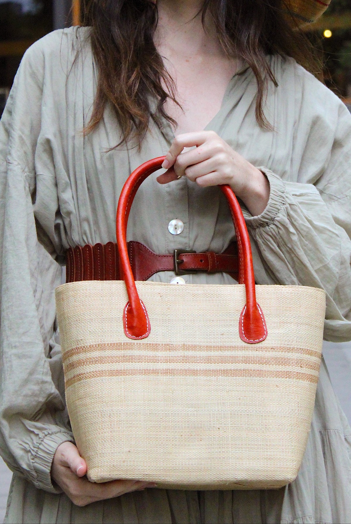 Model wearing Raja Racing Stripe Straw Basket Bag handmade loomed natural raffia palm fiber in a two tone multicolor design of Natural straw color with three horizontal bands of Cappuccino tea/light/brown creating a racing stripe pattern placed under the base of the handles a third down the height of the bag - Shebobo (with Monica 5" & 7" Wide Brim Multicolor Pinstripe Pattern Packable Straw Sun Hats)