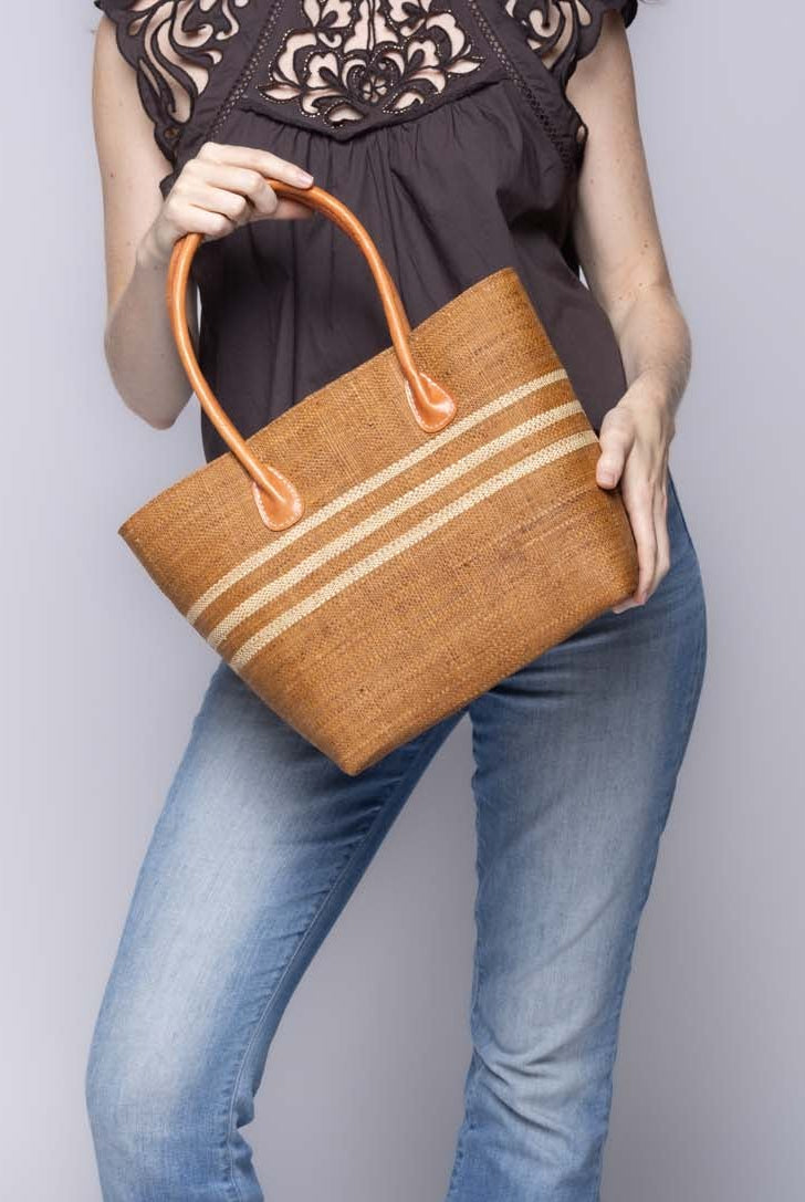 Model wearing Raja Racing Stripe Straw Basket Bag handmade loomed natural raffia palm fiber in a two tone multicolor design of Tobacco cinnamon/dark/brown color with three horizontal bands of Natural straw color creating a racing stripe pattern placed under the base of the handles a third down the height of the bag - Shebobo
