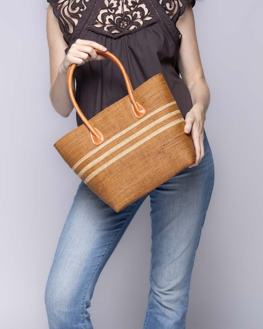 Model wearing Raja Racing Stripe Straw Basket Bag handmade loomed natural raffia palm fiber in a two tone multicolor design of Tobacco cinnamon/dark/brown color with three horizontal bands of Natural straw color creating a racing stripe pattern placed under the base of the handles a third down the height of the bag - Shebobo