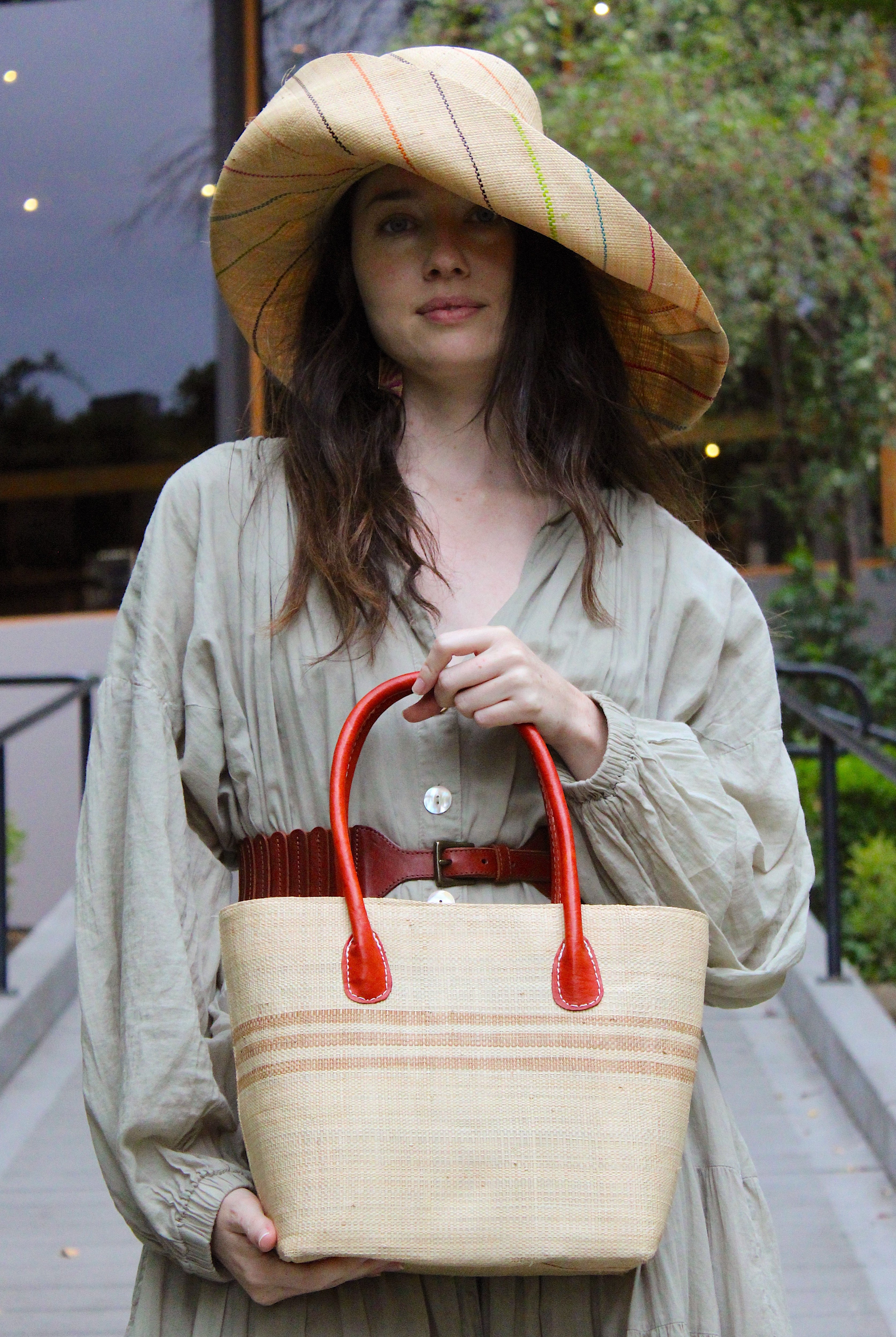 Model wearing Raja Racing Stripe Straw Basket Bag handmade loomed natural raffia palm fiber in a two tone multicolor design of Natural straw color with three horizontal bands of Cappuccino tea/light/brown creating a racing stripe pattern placed under the base of the handles a third down the height of the bag - Shebobo (with Monica 5" & 7" Wide Brim Multicolor Pinstripe Pattern Packable Straw Sun Hats)