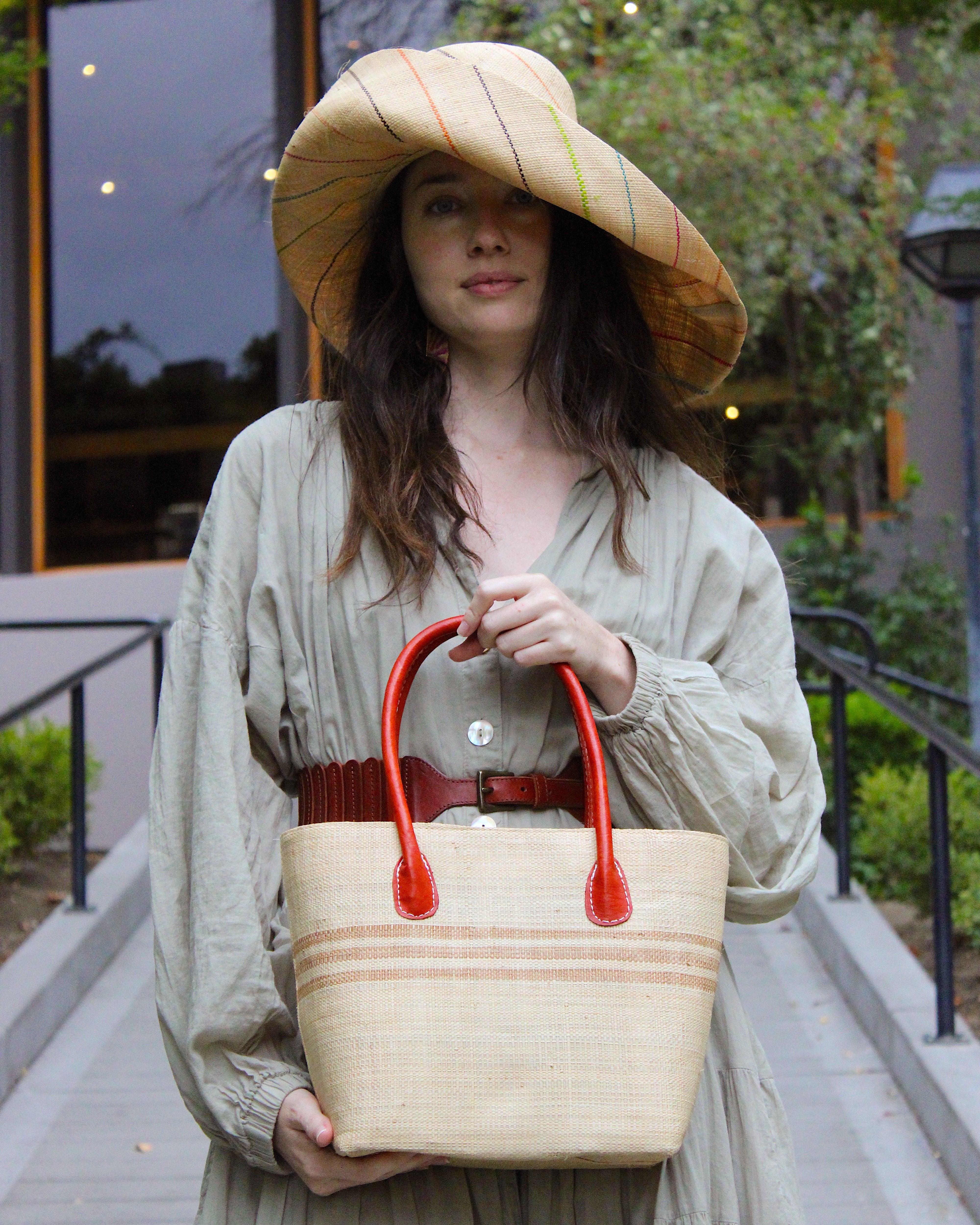Model wearing Raja Racing Stripe Straw Basket Bag handmade loomed natural raffia palm fiber in a two tone multicolor design of Natural straw color with three horizontal bands of Cappuccino tea/light/brown creating a racing stripe pattern placed under the base of the handles a third down the height of the bag - Shebobo (with Monica 5" & 7" Wide Brim Multicolor Pinstripe Pattern Packable Straw Sun Hats)