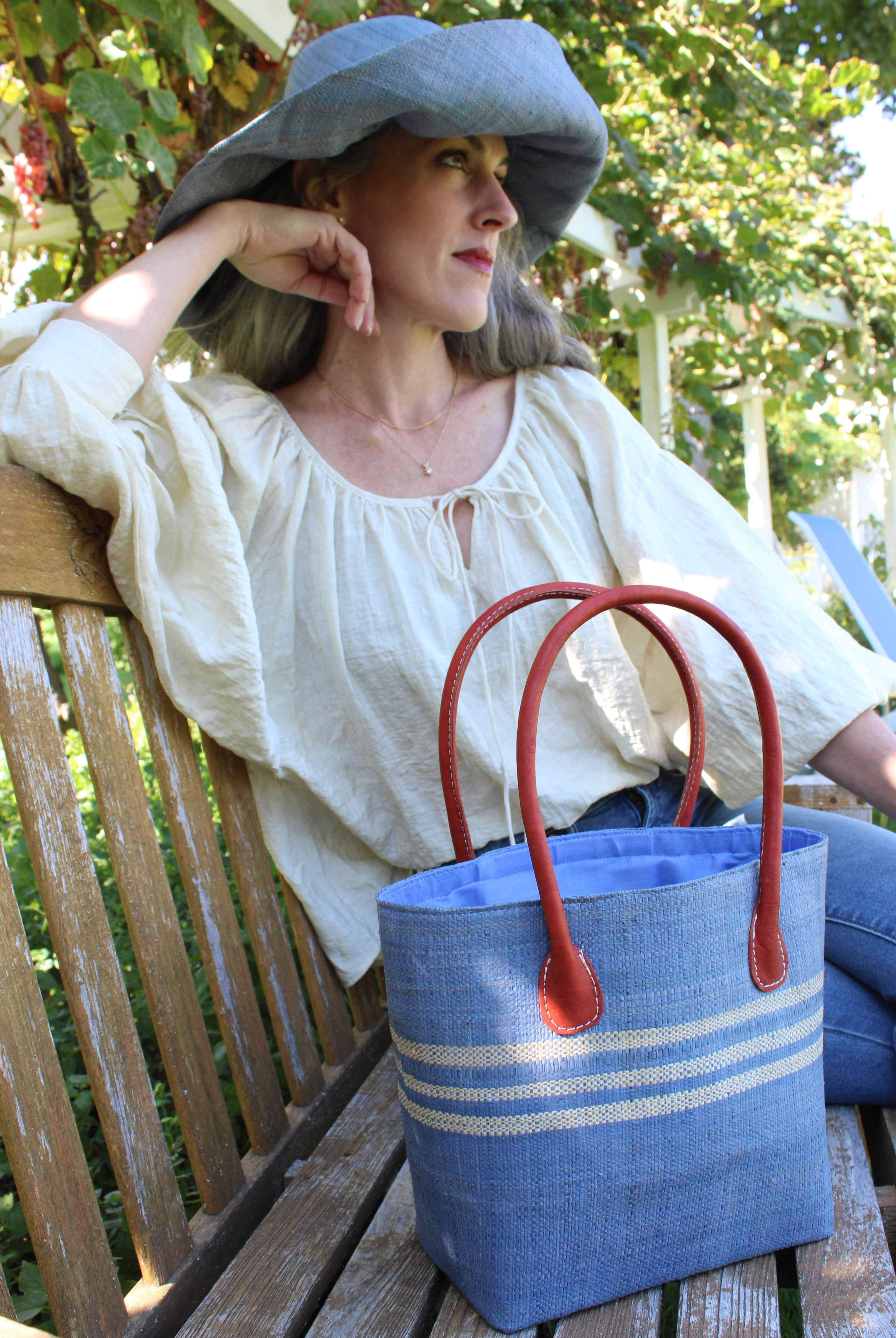 Model wearing Raja Racing Stripe Straw Basket Bag handmade loomed natural raffia palm fiber in a two tone multicolor design of Denim light/retro/soft/blue color with three horizontal bands of Natural straw color creating a racing stripe pattern placed under the base of the handles a third down the height of the bag - Shebobo (with Monica 5" & 7" Wide Brim Solid Color Packable Straw Sun Hats)