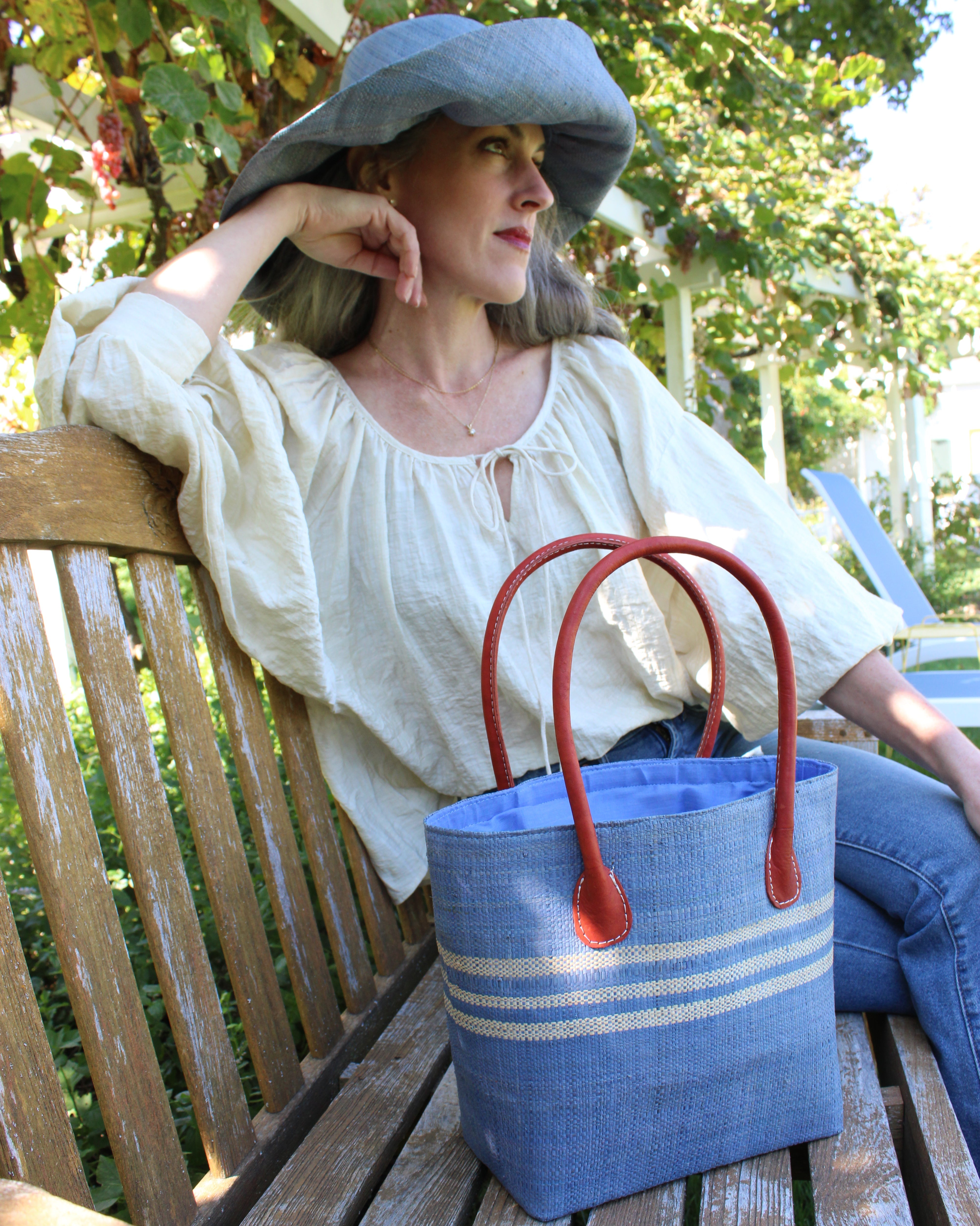 Model wearing Raja Racing Stripe Straw Basket Bag handmade loomed natural raffia palm fiber in a two tone multicolor design of Denim light/retro/soft/blue color with three horizontal bands of Natural straw color creating a racing stripe pattern placed under the base of the handles a third down the height of the bag - Shebobo (with Monica 5" & 7" Wide Brim Solid Color Packable Straw Sun Hats)