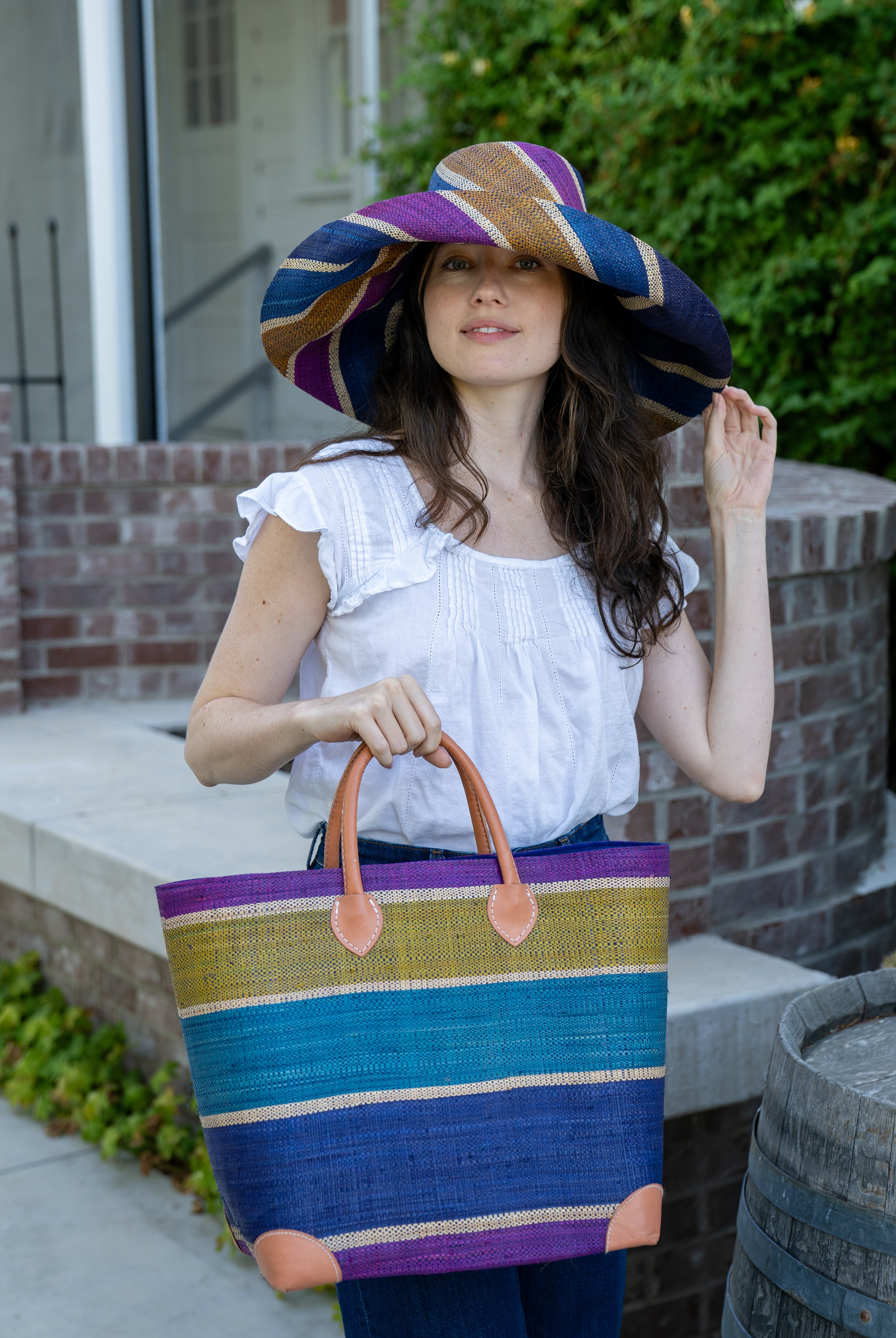 Model wearing Rayo Stripes Straw Basket Bag Olive Purple Multicolor navy blue, turquoise blue, olive green, purple, and natural straw color multiple width horizontal stripe pattern handmade loomed natural raffia palm fiber tote beach bag - Shebobo (with Monica 5" & 7" Wide Brim Multicolor Stripe Pattern Packable Straw Sun Hats)