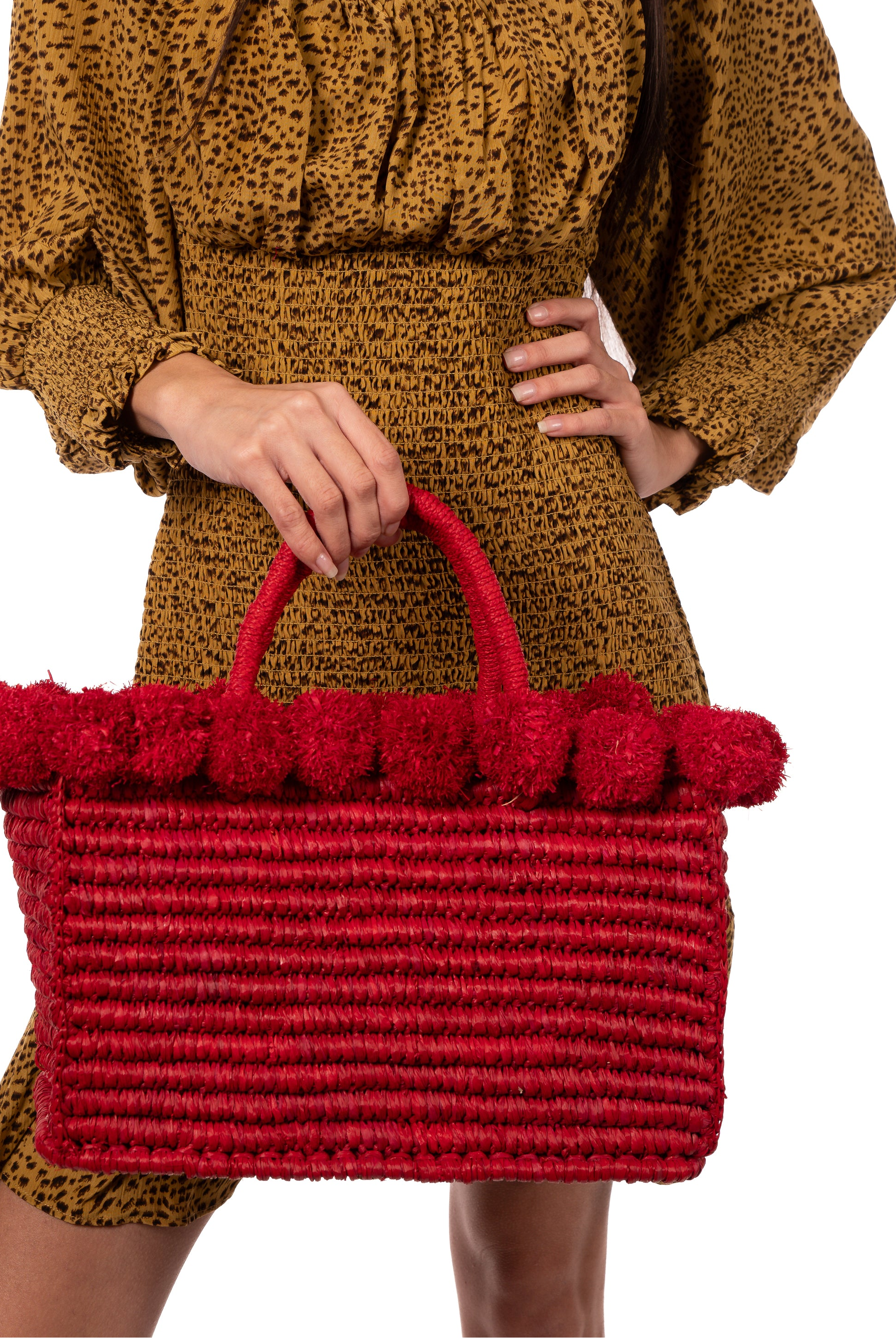 Model wearing Rico Red Crochet Rectangle Straw Basket handmade woven raffia palm fiber in a solid hue of red with horizontal bands of looped texture with matching small raffia pouf pompom top edge and wrapped braid handles small handbag purse - Shebobo