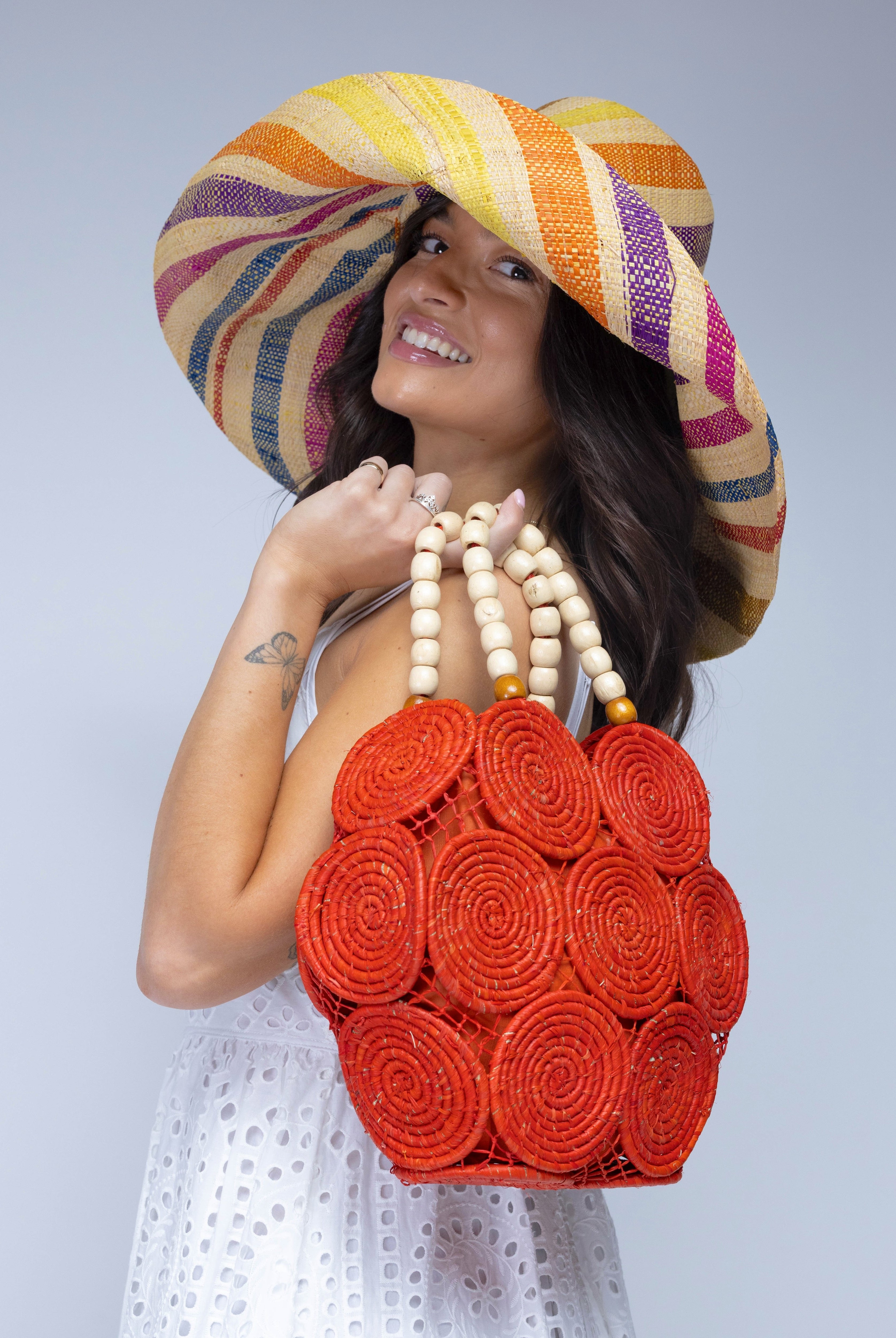 Model wearing Romy Coral Crochet Disc Straw Handbag with Wooden Bead Handles handmade natural raffia palm fiber in a solid hue of coral orange/red in a crochet coiled circular flat disc attached together with macrame knot technique with a geometric hexagonal overall shape with seven discs per side two above and below a horizontal row of three per side and a single row comprising the side of the bag with beige carved round wooden beaded handles purse - Shebobo (with Monica 7" Wide Brim Packable Straw Sun Hat