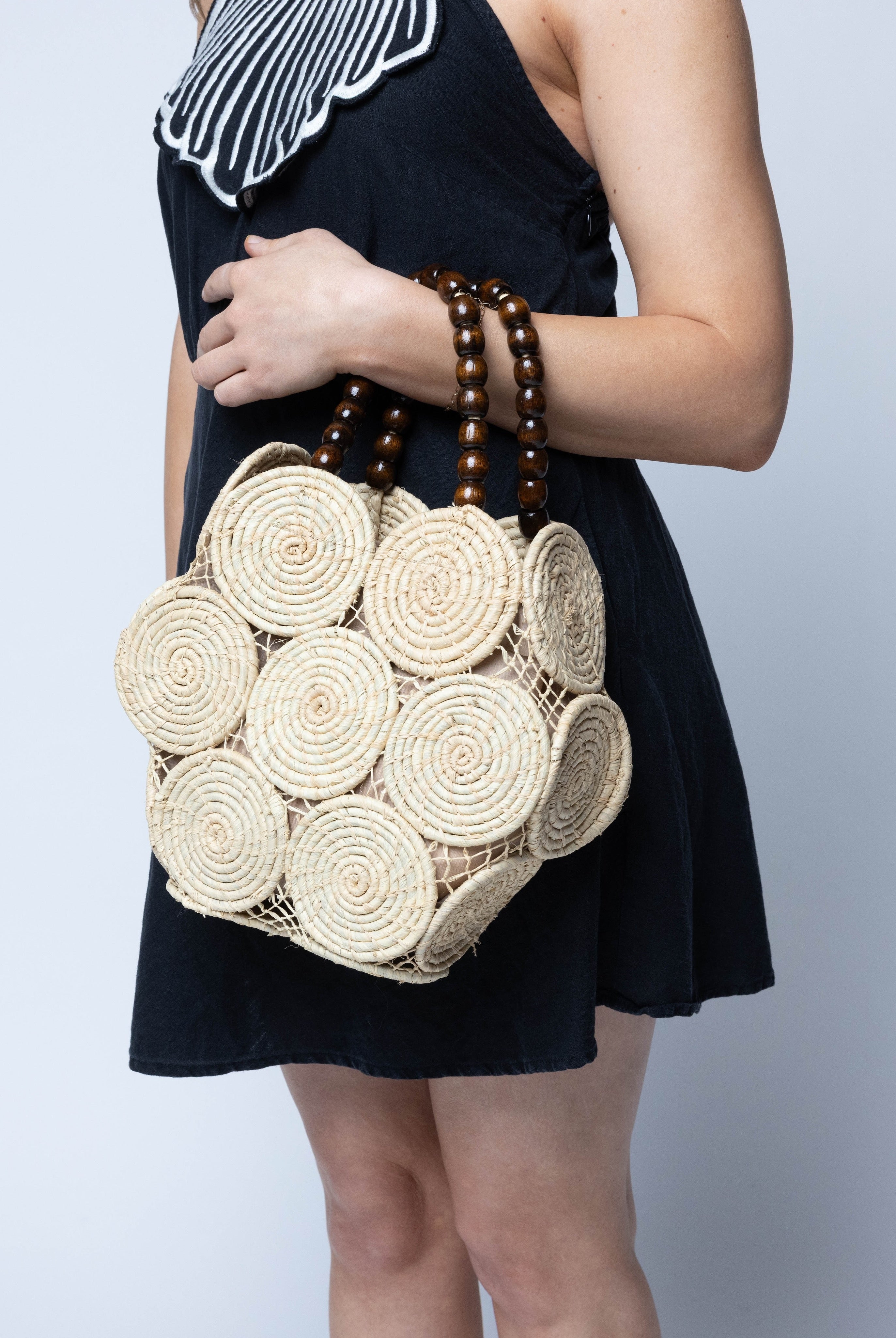 Model wearing Romy Natural Crochet Disc Straw Handbag with Wooden Bead Handles handmade natural raffia palm fiber in a solid hue of natural straw color in a crochet coiled circular flat disc attached together with macrame knot technique with a geometric hexagonal overall shape with seven discs per side two above and below a horizontal row of three per side and a single row comprising the side of the bag with dark brown carved round wooden beaded handles purse - Shebobo